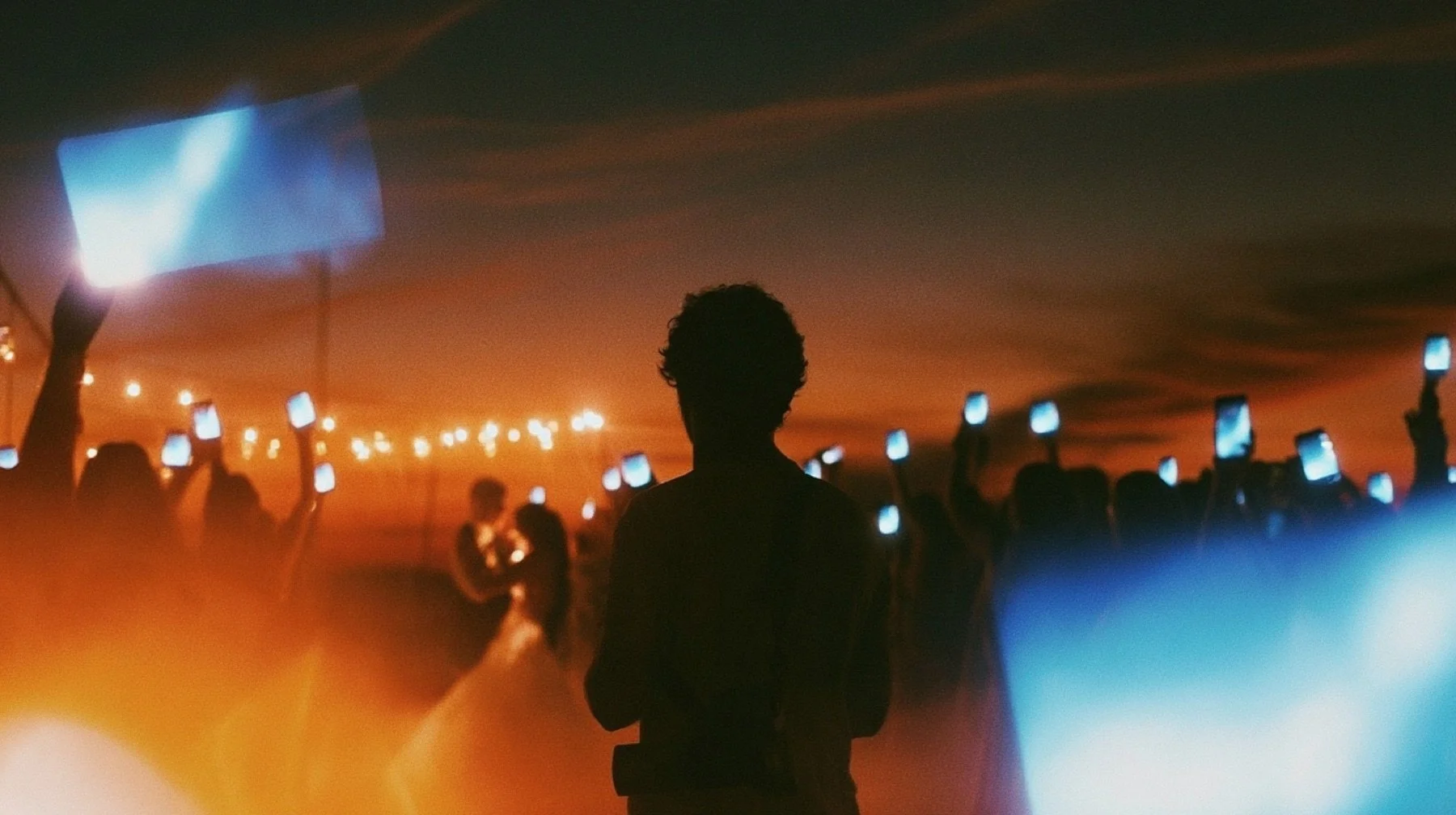 A wedding reception at dusk seen from behind a photographer with a camera hanging at his chest, dozens of guests holding up glowing phones toward the couple dancing under string lights.