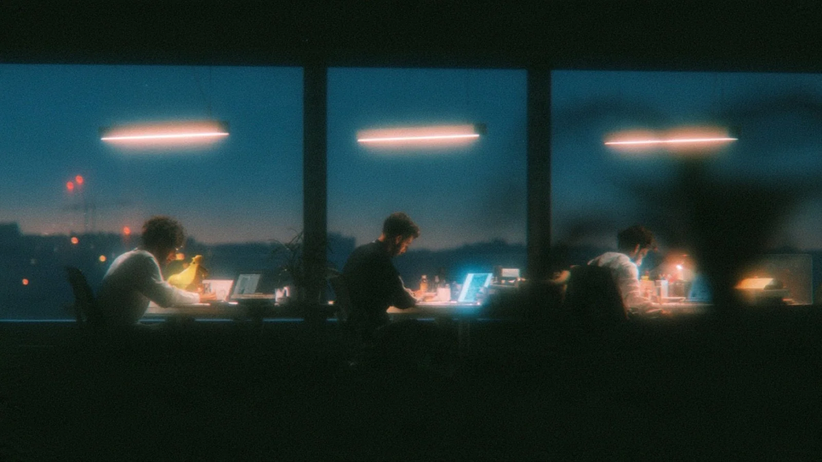 Three coworkers at three separate desks in one office at night, each in a different posture and none of them looking at each other.