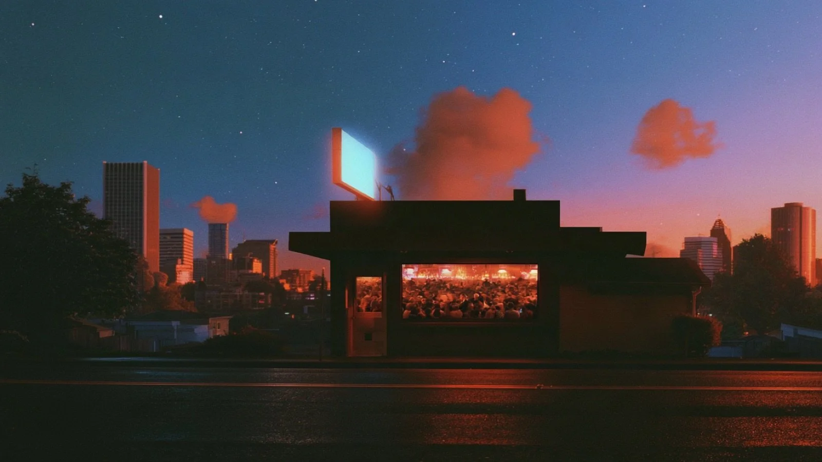 A small bar at dusk with a blank lit sign on its roof and a packed crowd visible through its front window, a city skyline behind it.