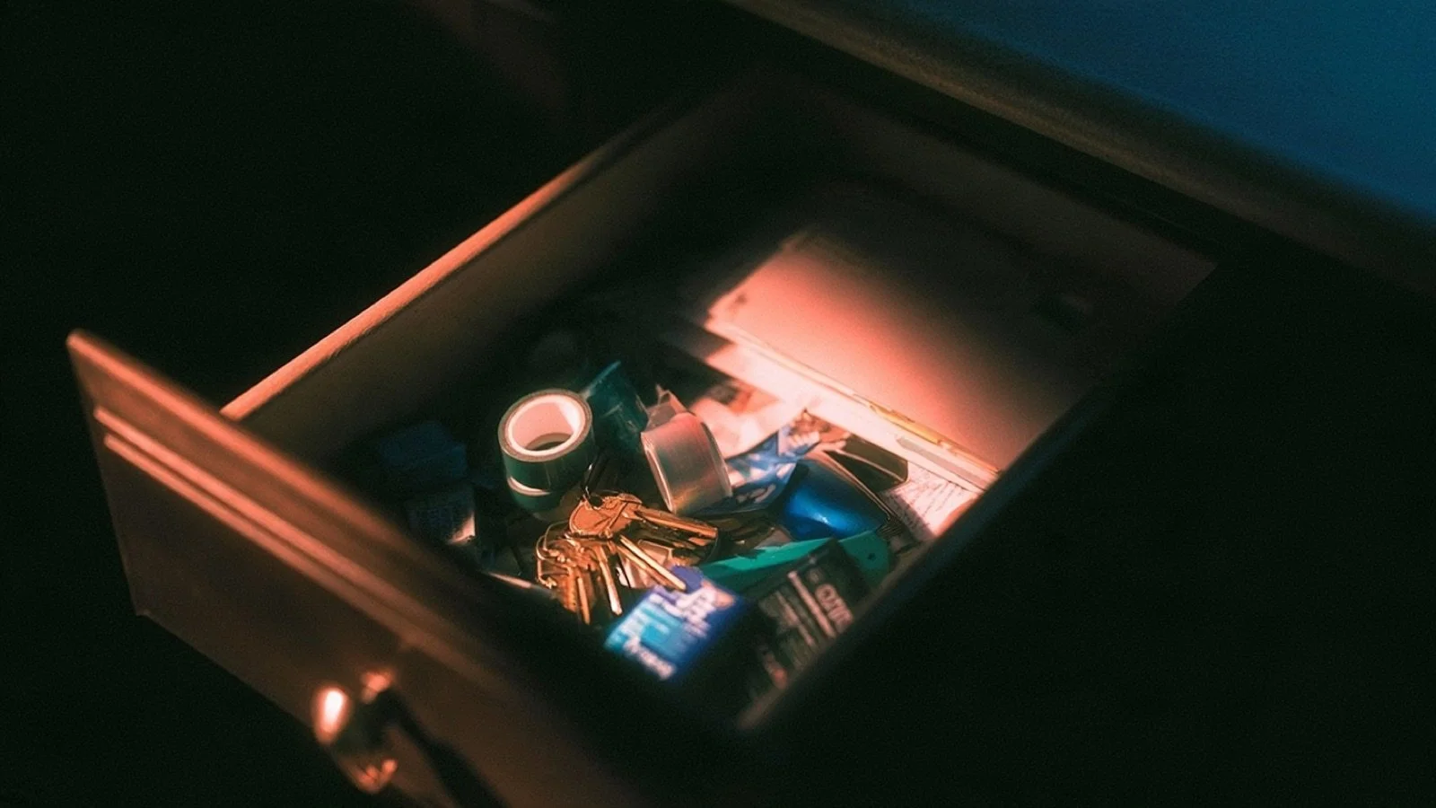 An open kitchen drawer seen from above, holding tape, loose keys, batteries and folded paperwork jumbled together in the warm light of a kitchen at evening.