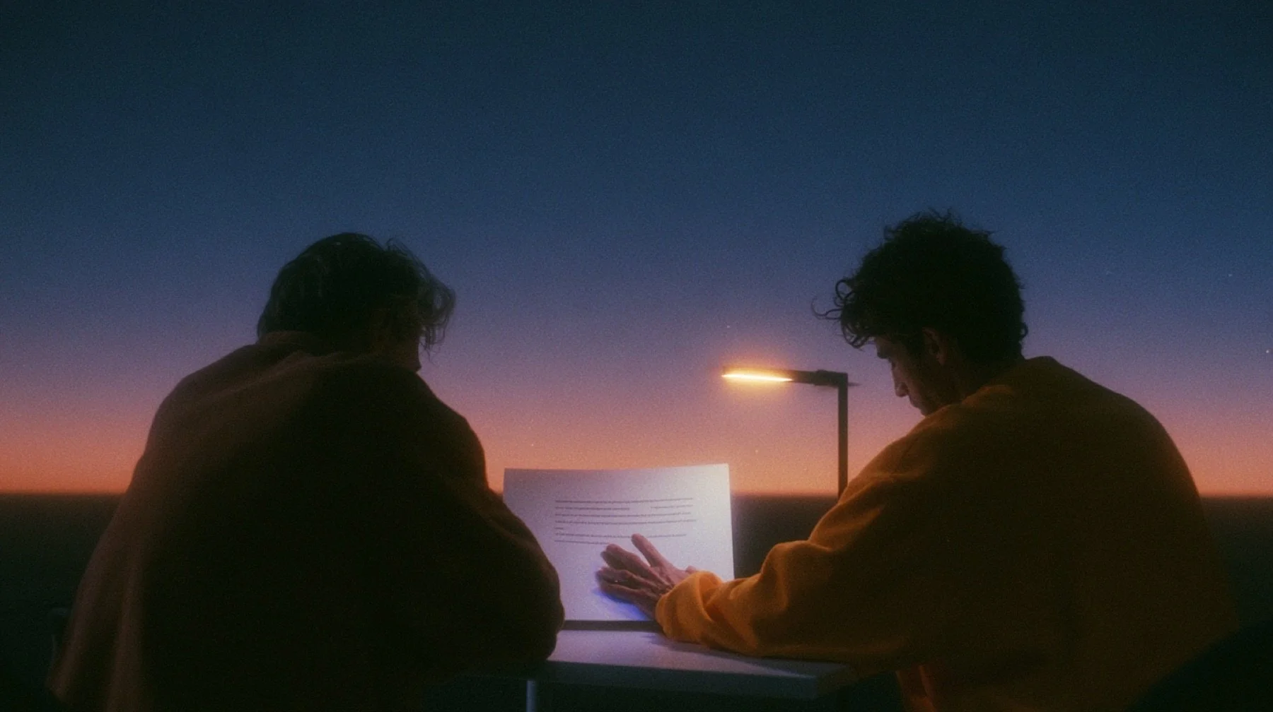 Two people seen from behind at a small desk at dusk, a single printed sheet between them and one hand laid flat on it.