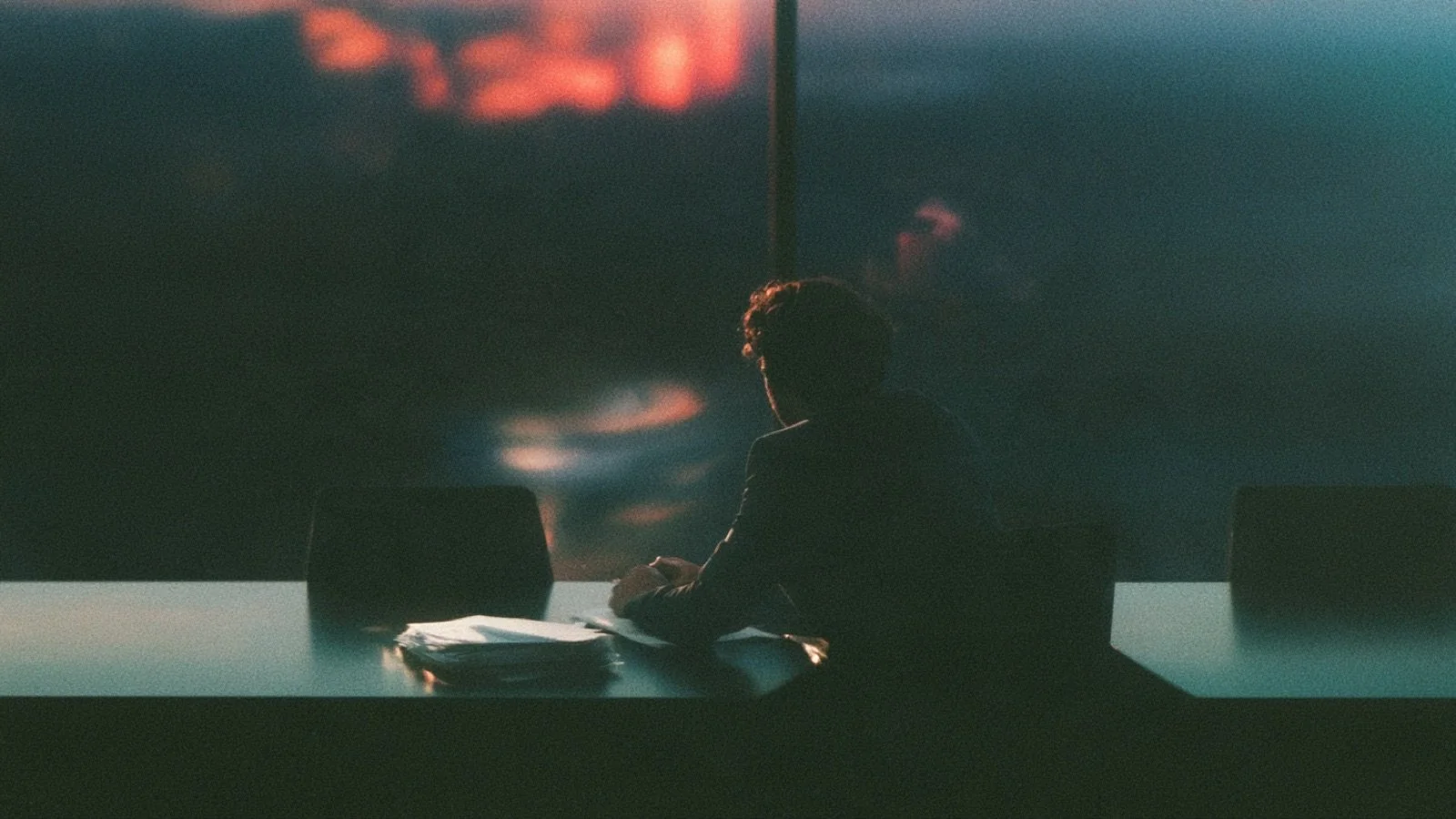 One person sitting alone at a long meeting table with a stack of papers in front of them and two empty chairs beside them at the same table.