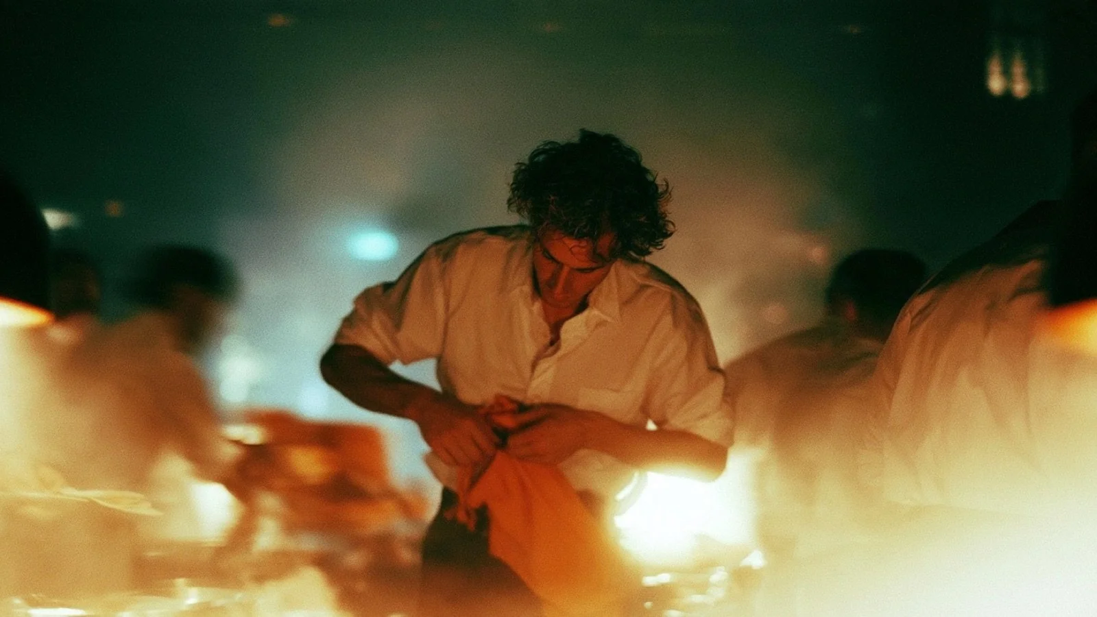 A cook in a white shirt holds an orange apron in a hot, steamy kitchen while other staff blur past him.