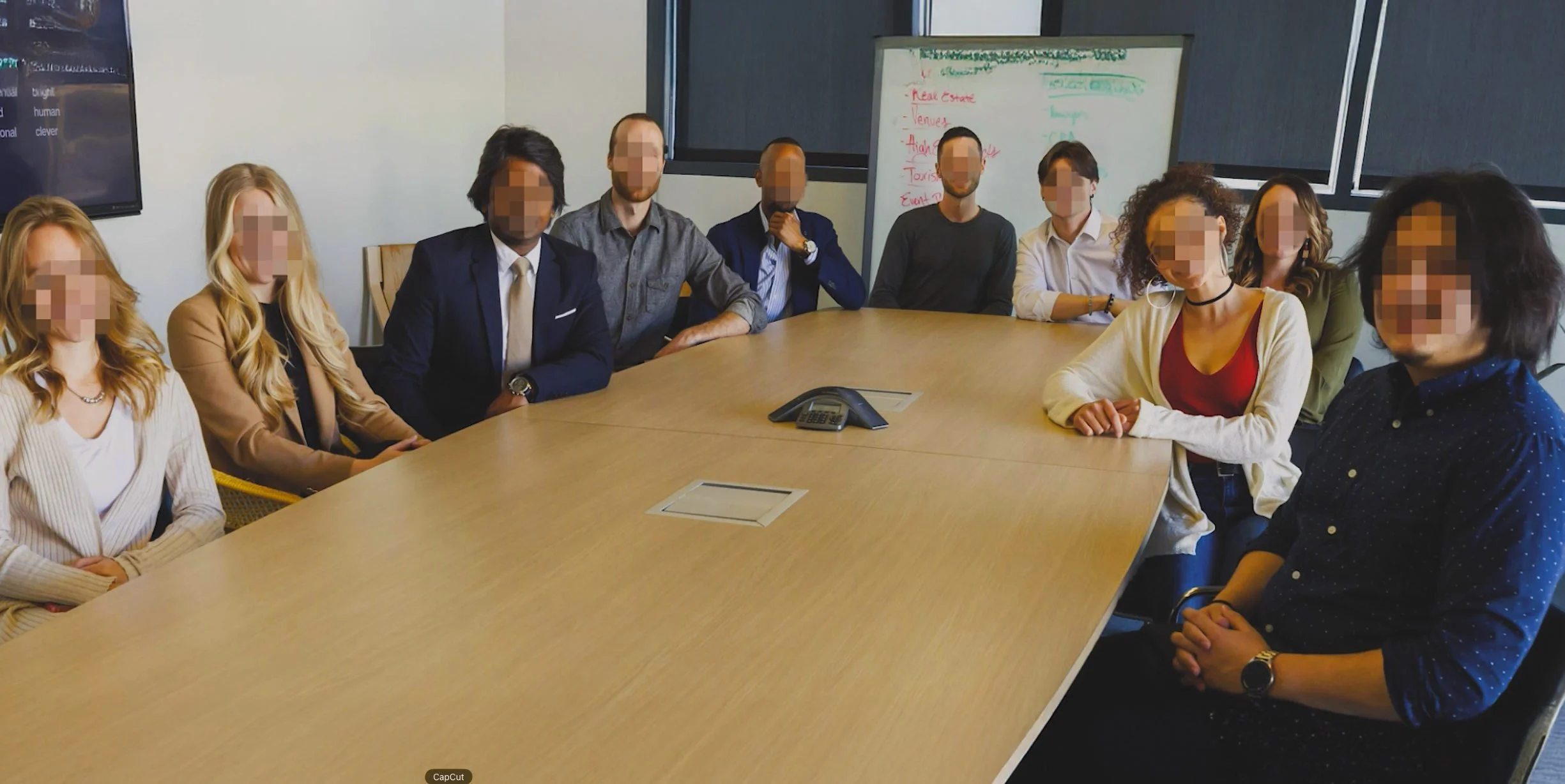 Group of diverse professionals sitting around a conference table in a meeting room, with whiteboard and screen in the background.