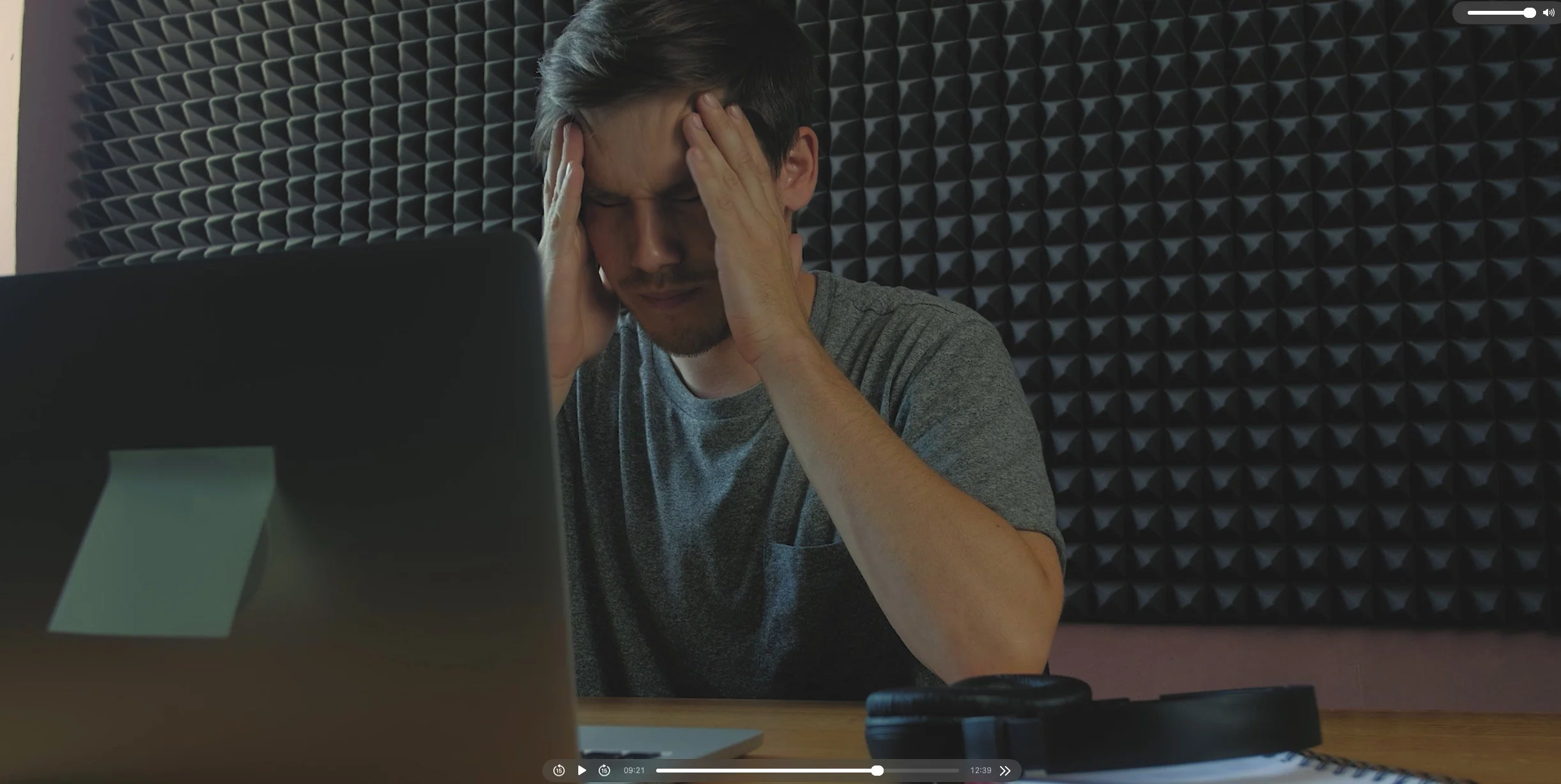 A young man sitting at a desk holding his head with both hands, appearing stressed or frustrated, with a laptop and headphones on the desk. The background features soundproofing foam panels.