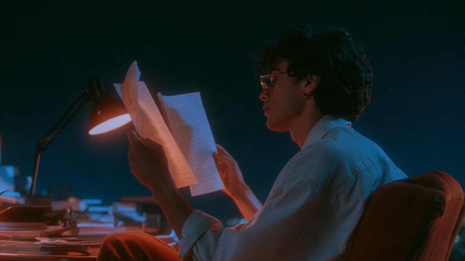 A person at a desk at night holding up two separate documents, one in each hand, comparing them under the warm light of a desk lamp.