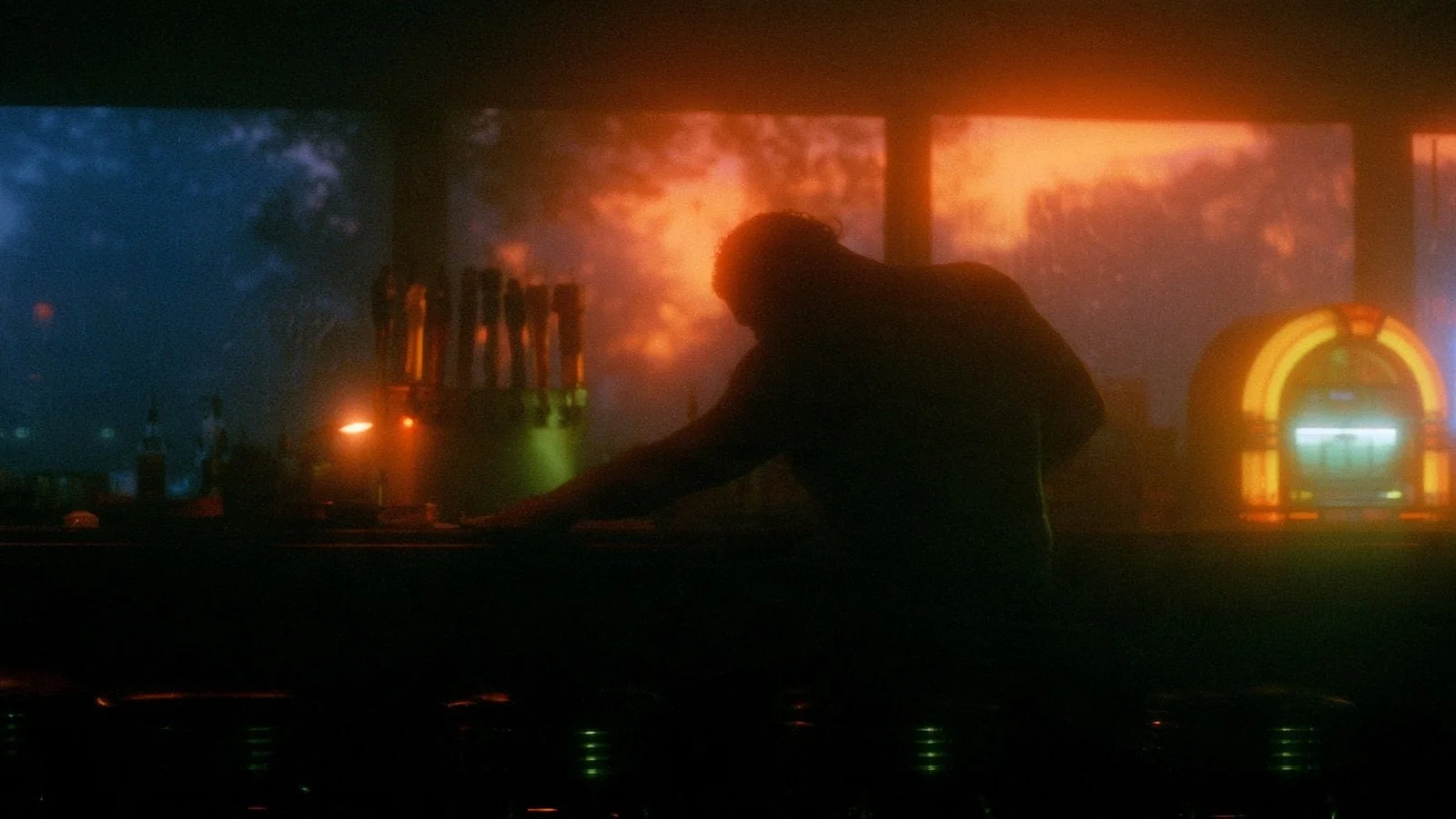 A lone bartender leans on the counter of an empty bar at evening, the beer taps and a glowing jukebox silhouetted against the windows.