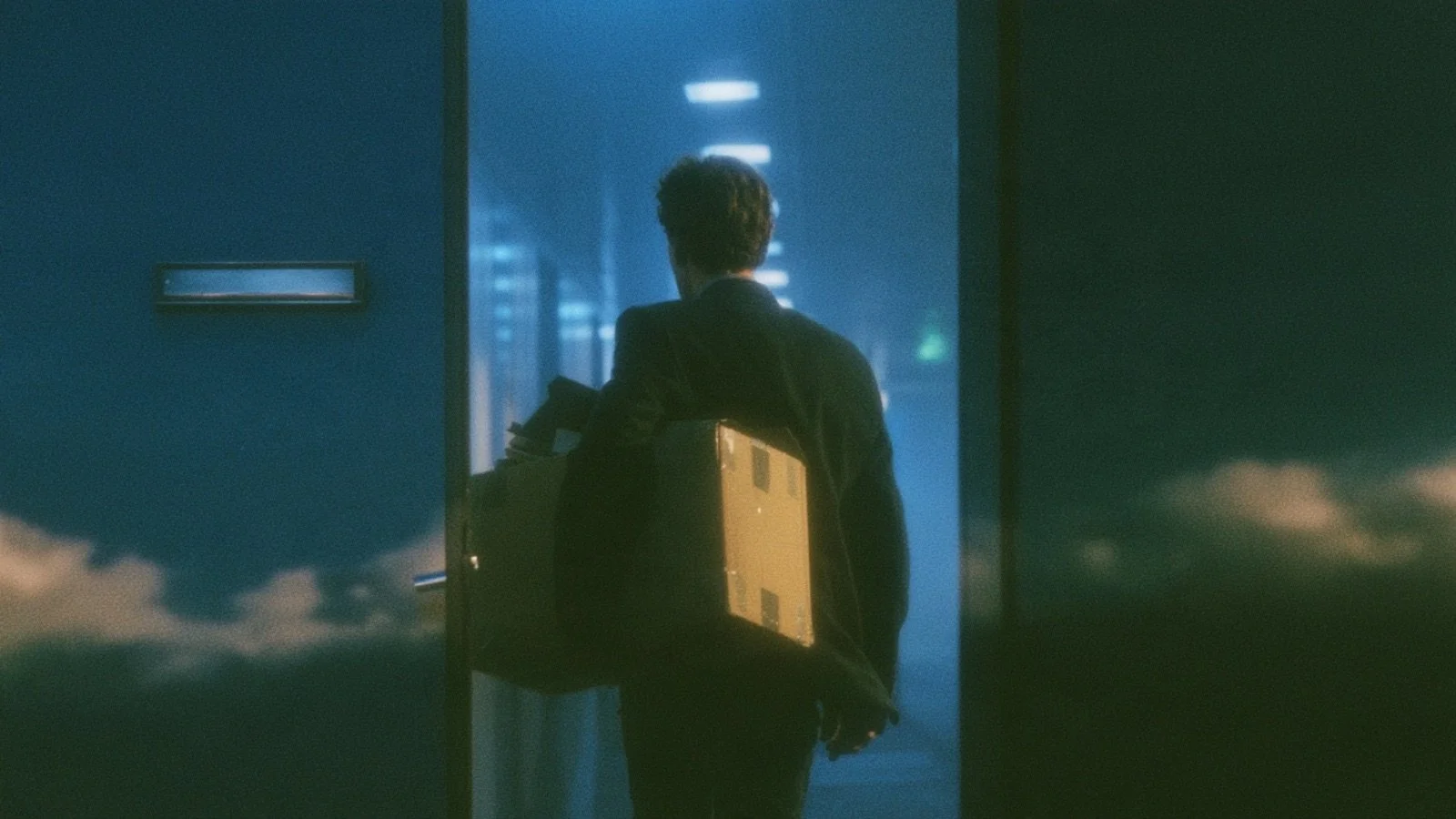 A person seen from behind carrying a cardboard box of belongings out through an office doorway, an empty nameplate holder on the wall beside them.