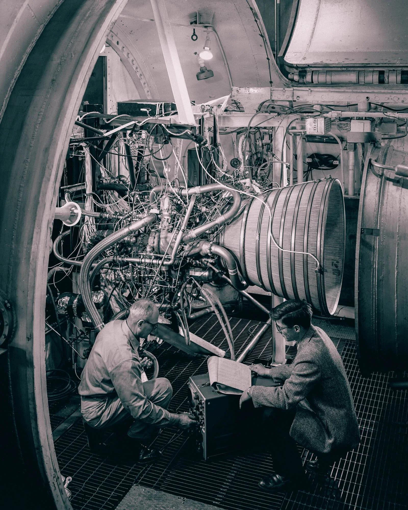 Two engineers crouched beside a rocket engine inside a test chamber, one of them taking notes