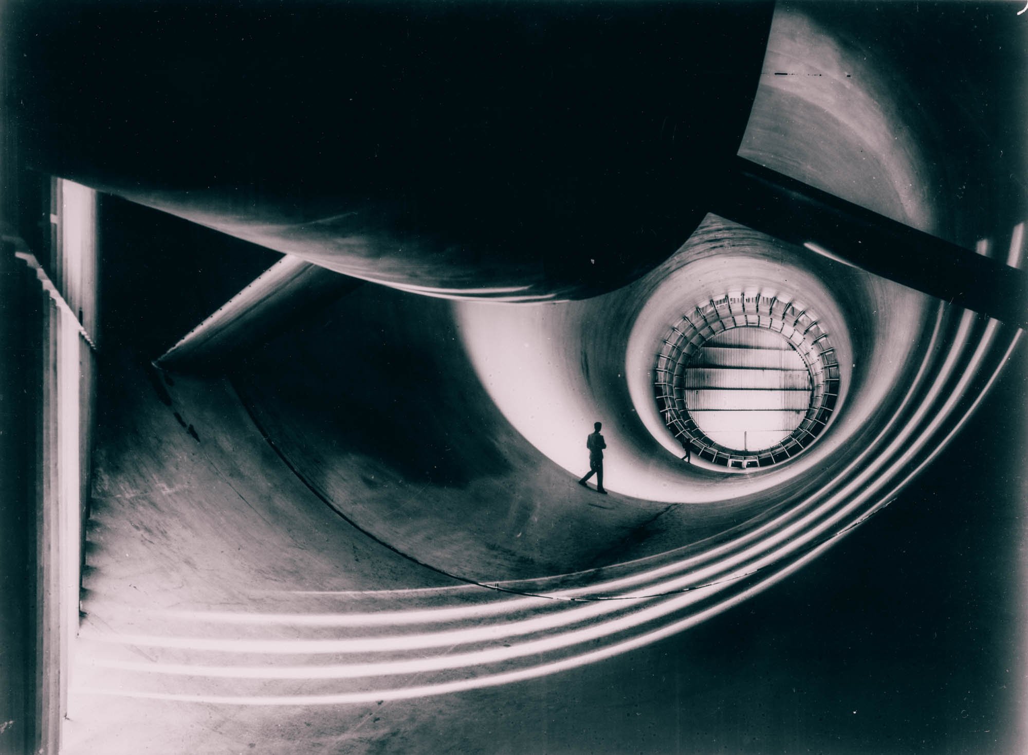 A lone figure standing inside the throat of an enormous wind tunnel, dwarfed by the curved walls
