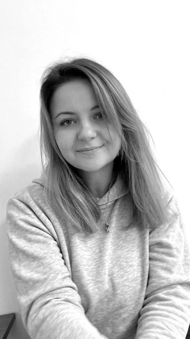 Black and white photo of a young woman with shoulder-length hair, smiling, wearing a hoodie and a necklace, sitting against a plain wall.
