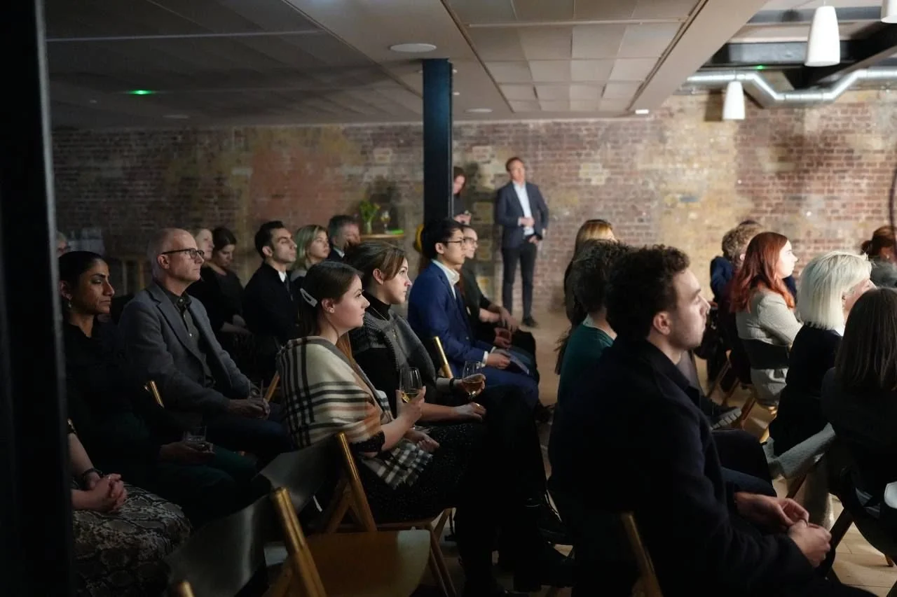 People attending a presentation or speech at an indoor event with a brick wall background, some seated and some standing in the back.