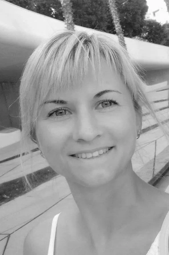 A smiling woman with short blond hair standing outdoors near a boat with trees in the background.