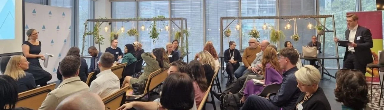 A conference room filled with diverse attendees seated and listening to a speaker standing at the front. The room has large windows, hanging plants, and a decorated backdrop.