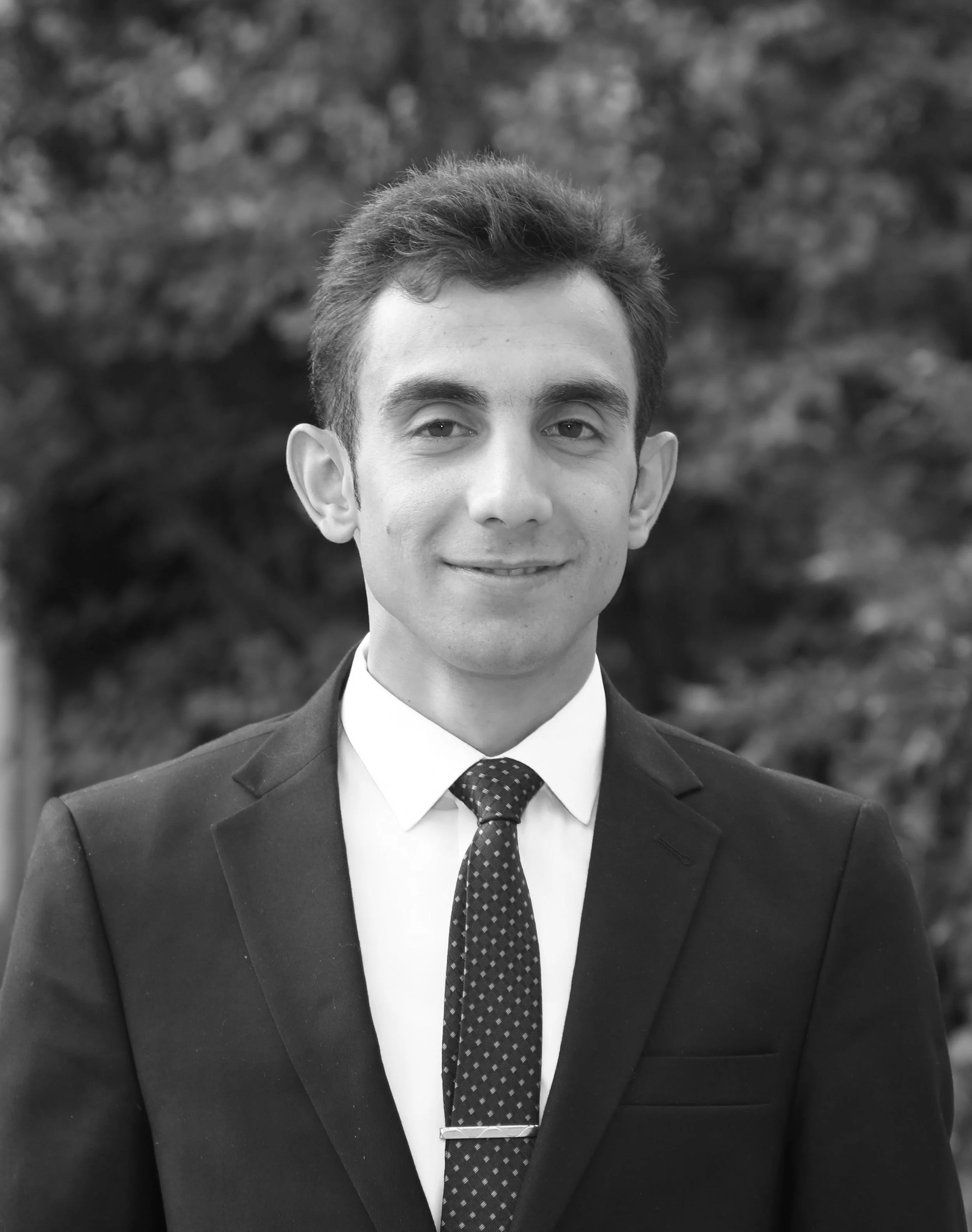 Black and white portrait of a young man in a suit and tie, smiling slightly, with a blurred outdoor background.
