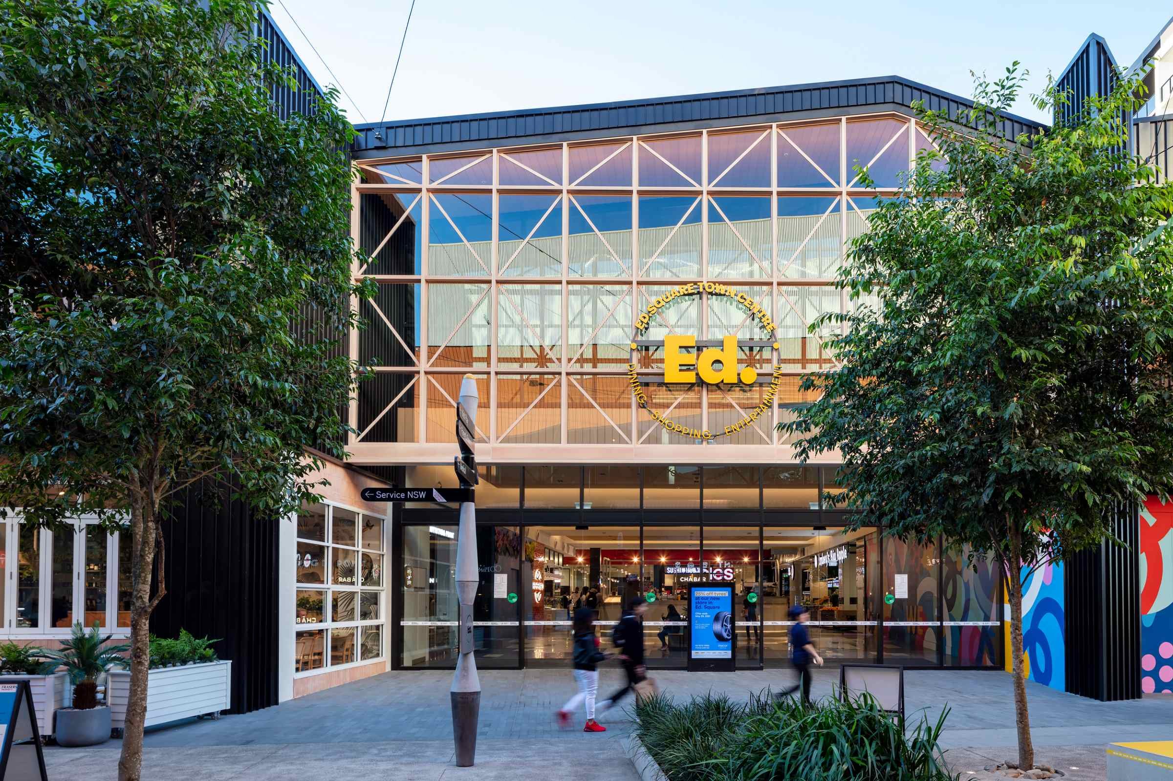 Exterior view of a shopping center entrance named Ed. with a large glass facade, trees on either side, and people walking in front during daytime.