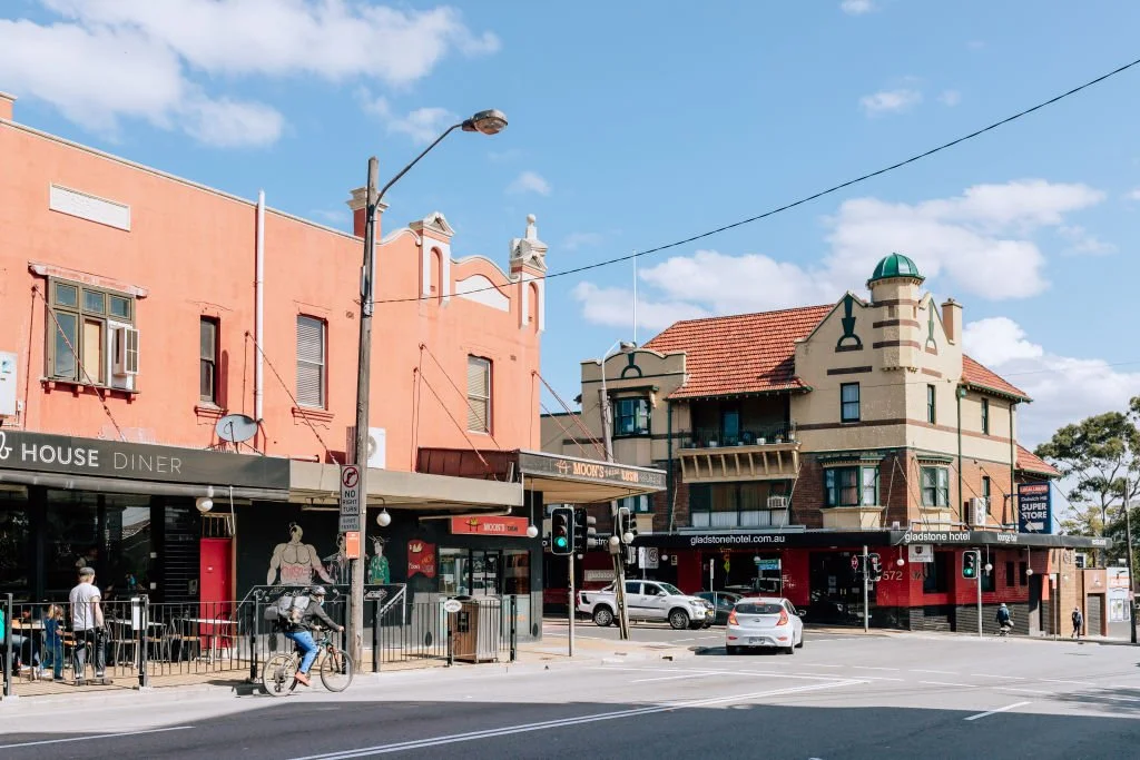 City street scene with buildings, cars, a cyclist, and pedestrians on the sidewalk on a sunny day.