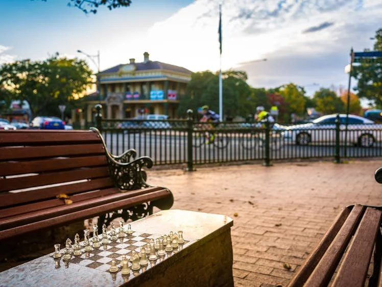 Outdoor scene with benches, a chessboard, and a wrought iron fence in front of a two-story building with balconies, trees, and parked cars.