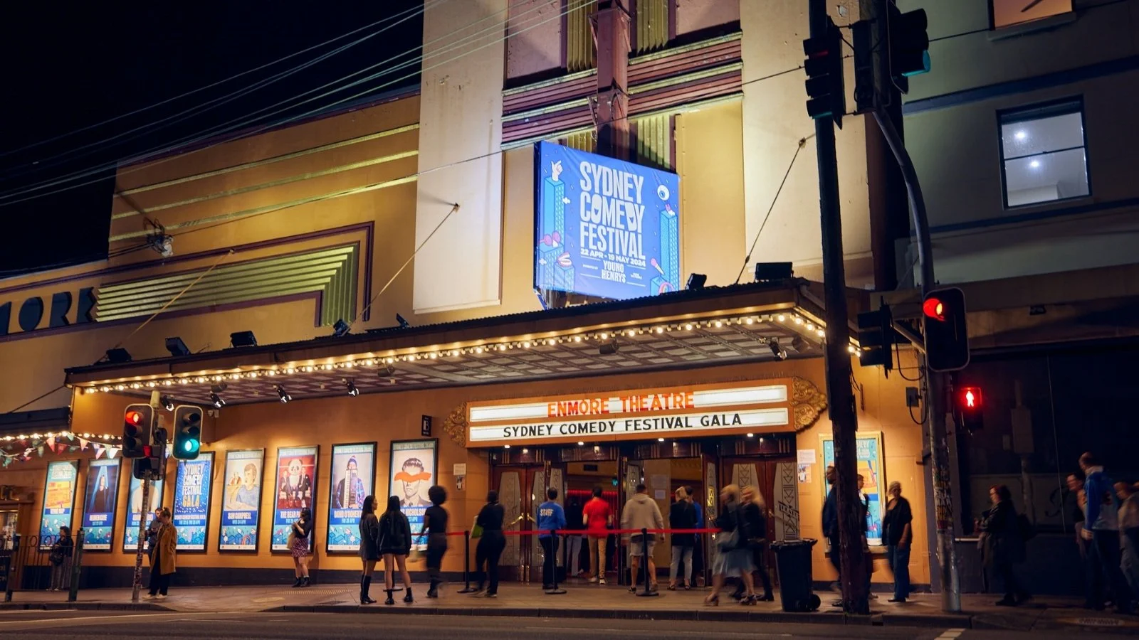 Night view of a theatre marquee for Enmore Theatre with a line of people waiting outside, promoting the Sydney Comedy Festival Gala, with bright lights, posters, and a digital sign overhead.