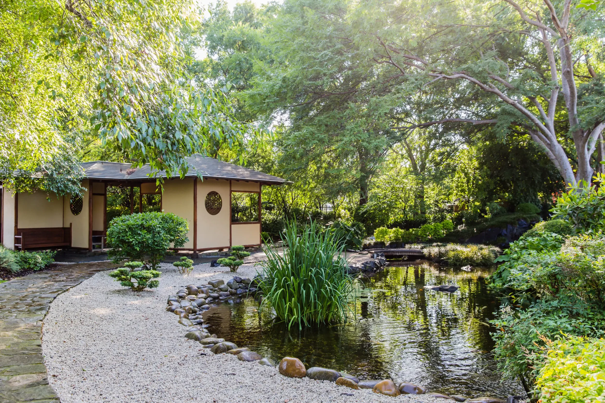 A serene Japanese-style garden with a small pond, stone pathway, lush green trees, and a traditional pavilion in the background.