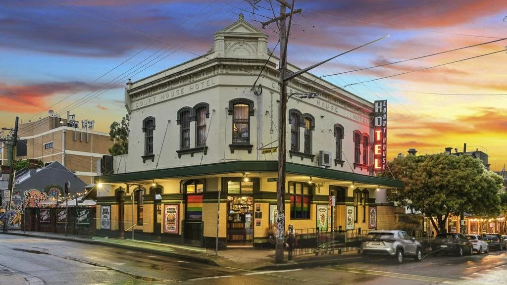A two-story hotel building on a street corner during sunset, with a neon sign reading 'Hotel' and colorful pride flags displayed outside.