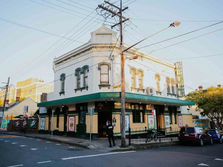 A two-story corner building with a green awning, street signs, parked car, power lines, and people outside in an urban area during sunset.