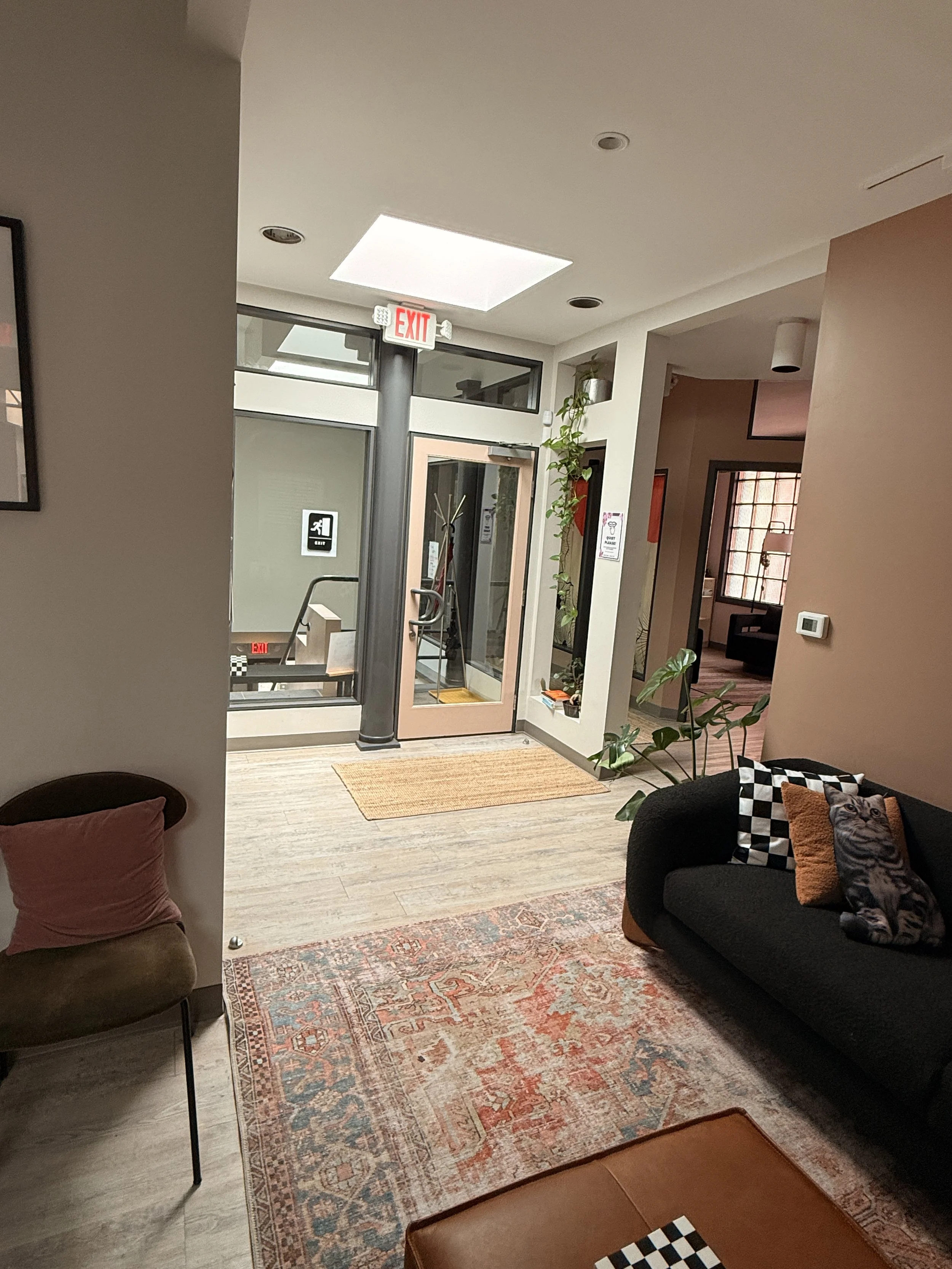 Interior of a modern apartment or hotel lobby with a black sofa, patterned cushions, a plant, a rug, and a glass door leading outside, with an exit sign and ceiling lights.