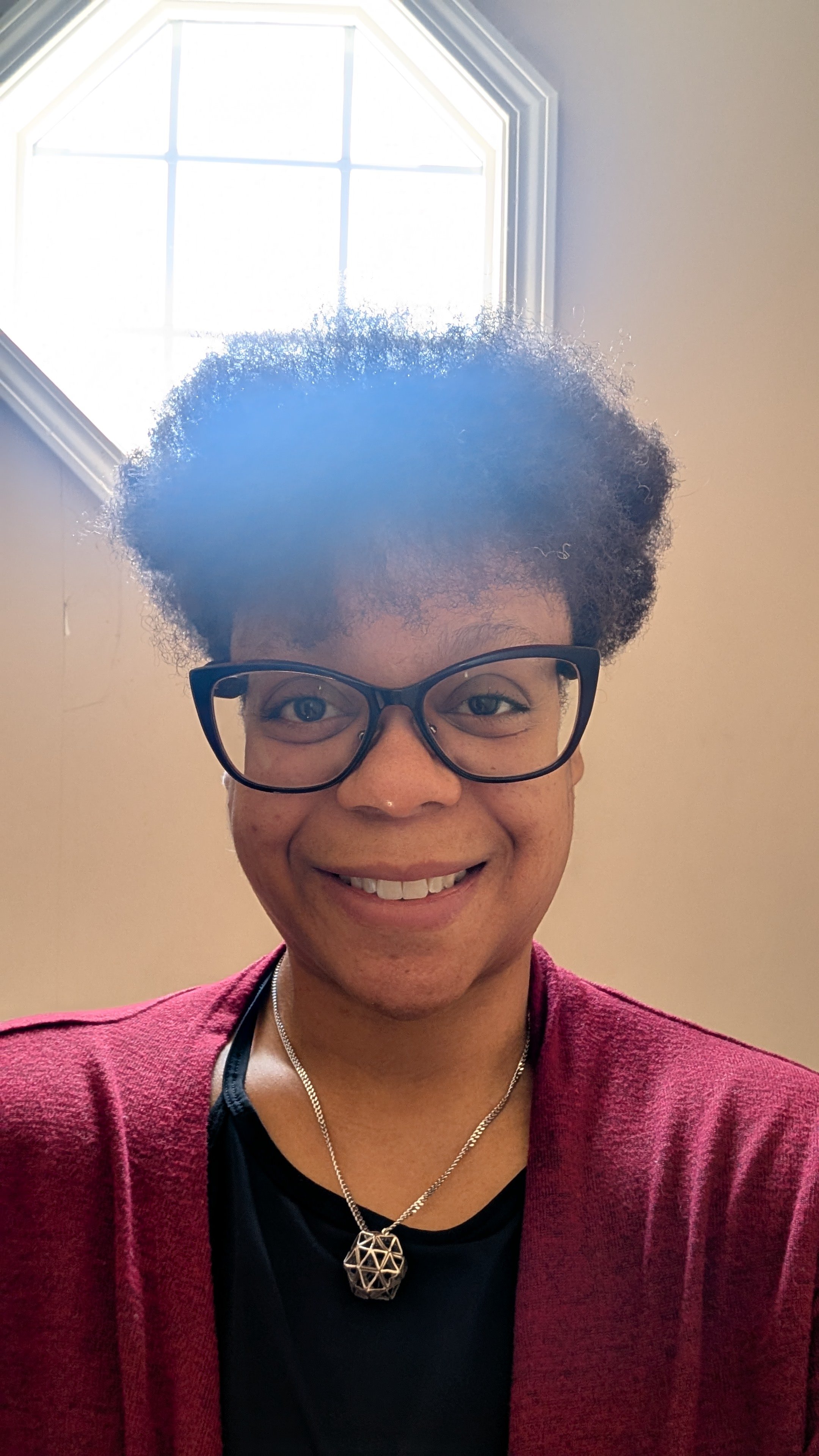 Person with glasses with curly hair smiling in front of a window.