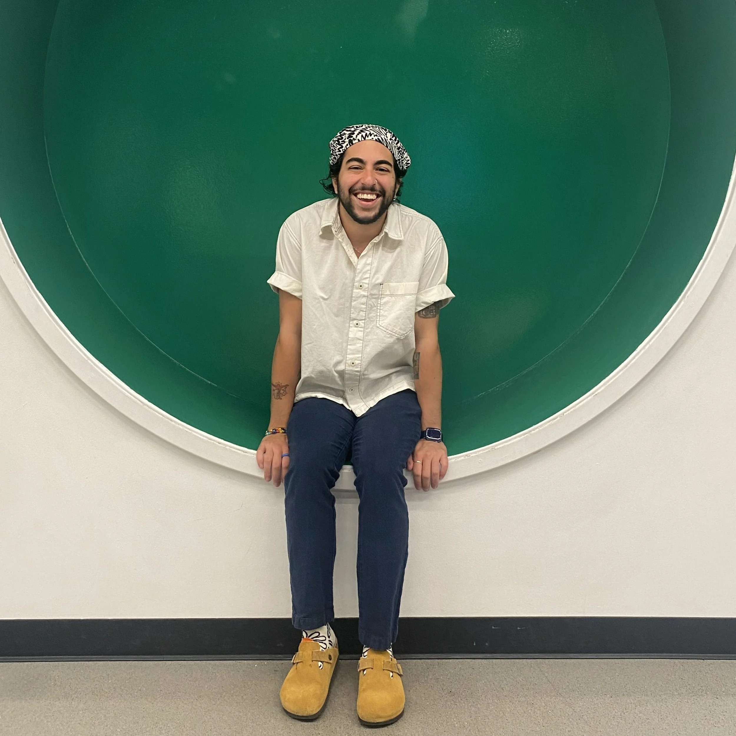 Person with dark curly hair in a bandana, a dark beard, and tan skin smiles at the camera while sitting at the edge of a circular alcove.