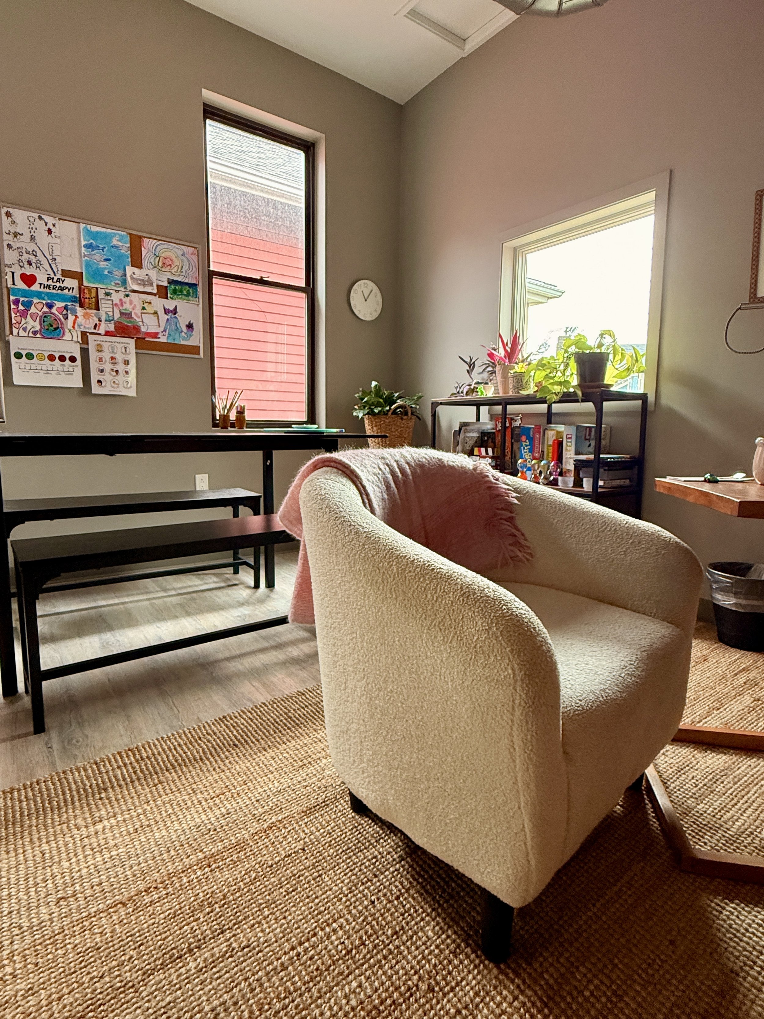 A cozy room with a white armchair on a woven rug, a wall with colorful children's artwork, and a window with plants. We have several clinicians who specialize in child and teen therapy. 