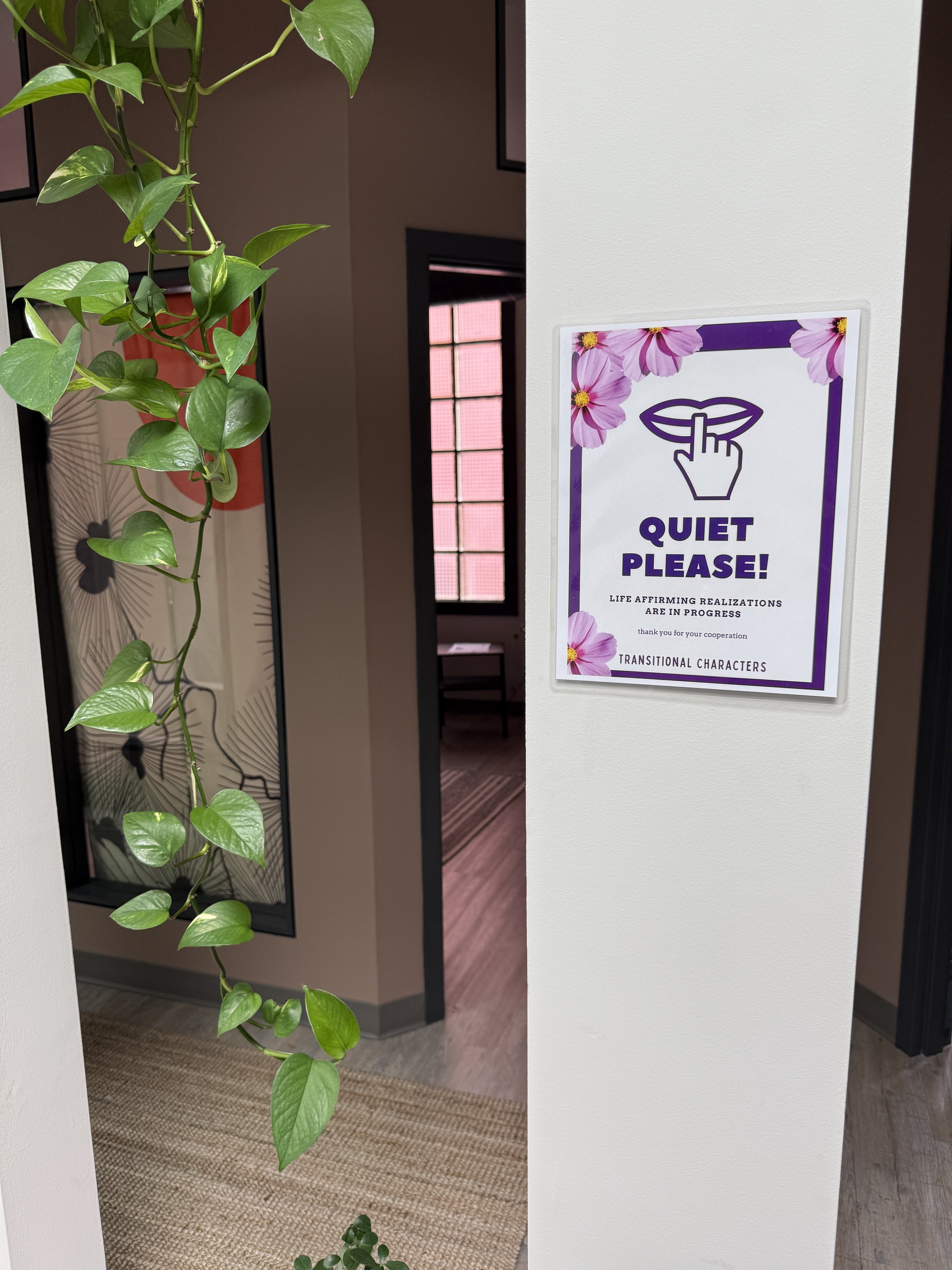 A sign on a white wall reads 'Quiet Please!' with a graphic of a hand with the index finger over lips, surrounded by purple and pink flowers. A green leafy plant is hanging nearby.