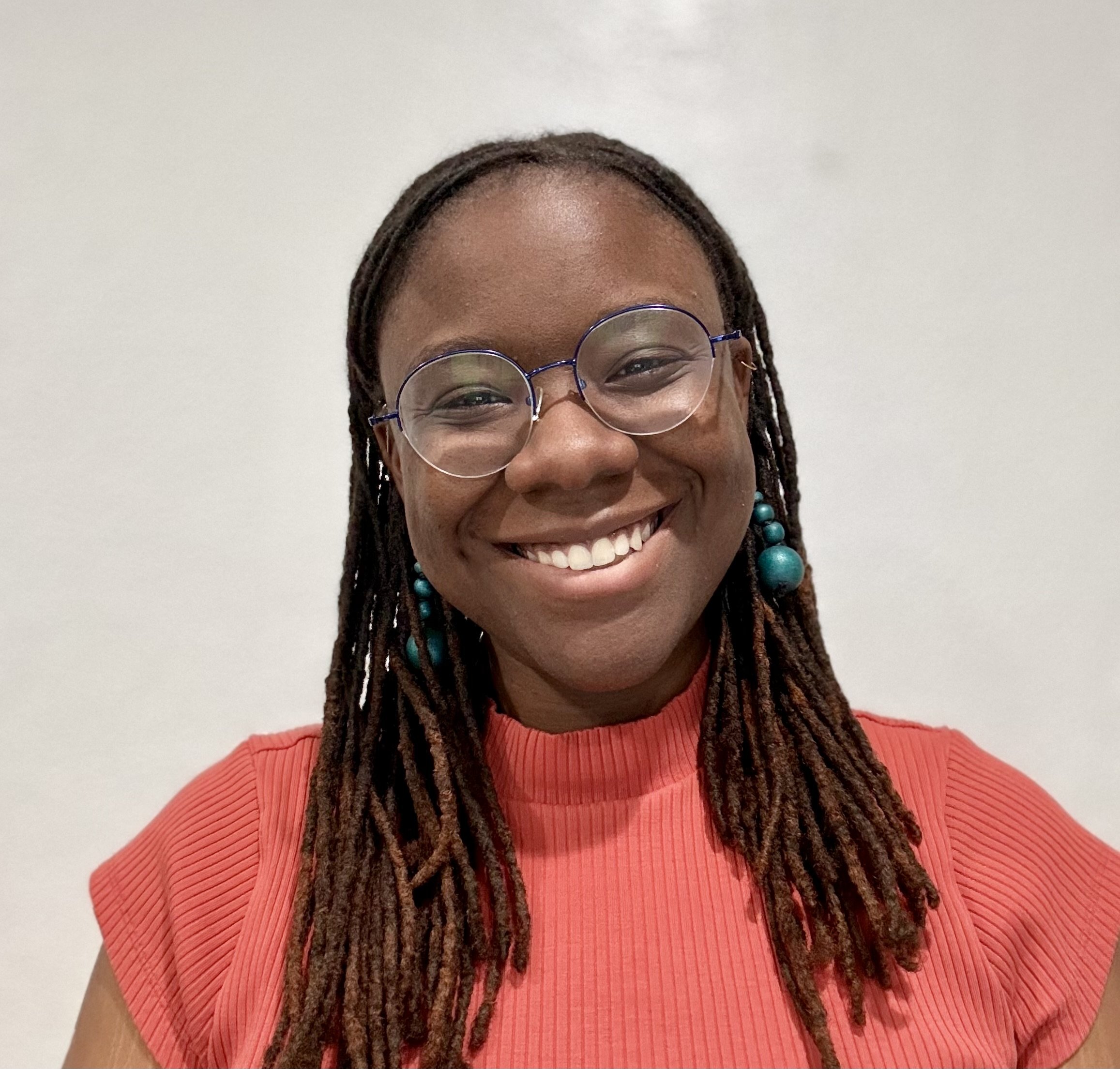 Person smiling into the camera in a pink shirt, with locs and a blue dangling earrings.