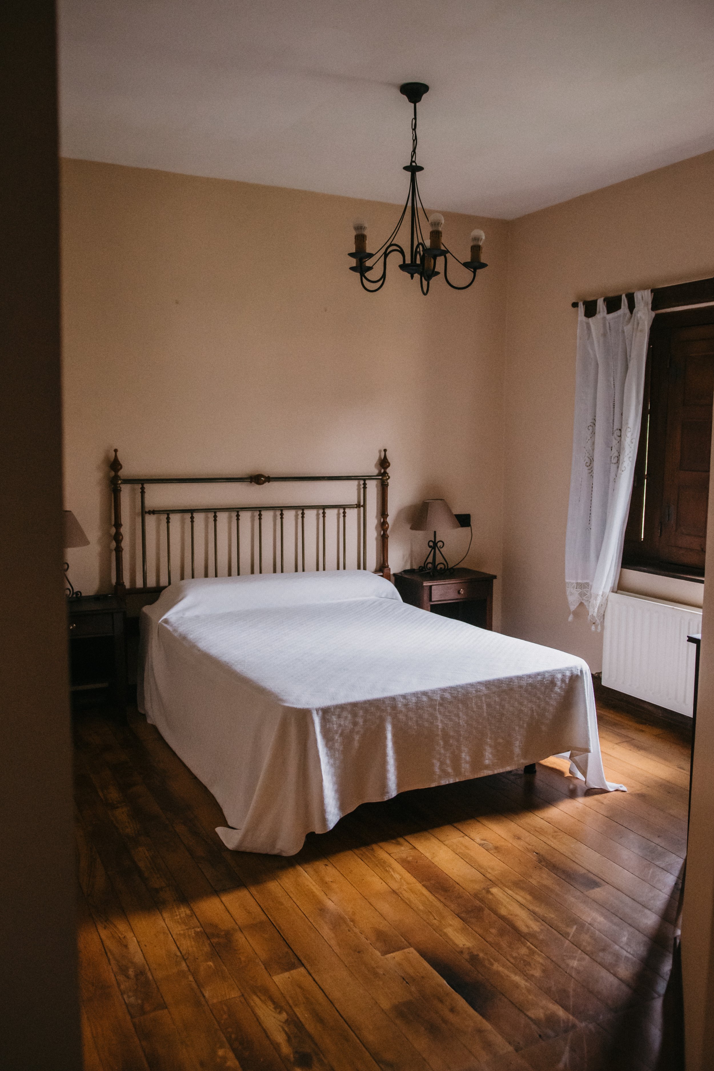 Habitación con cama cubierta con sábanas blancas, dos mesas de noche con lámparas y una ventana con cortinas blancas.