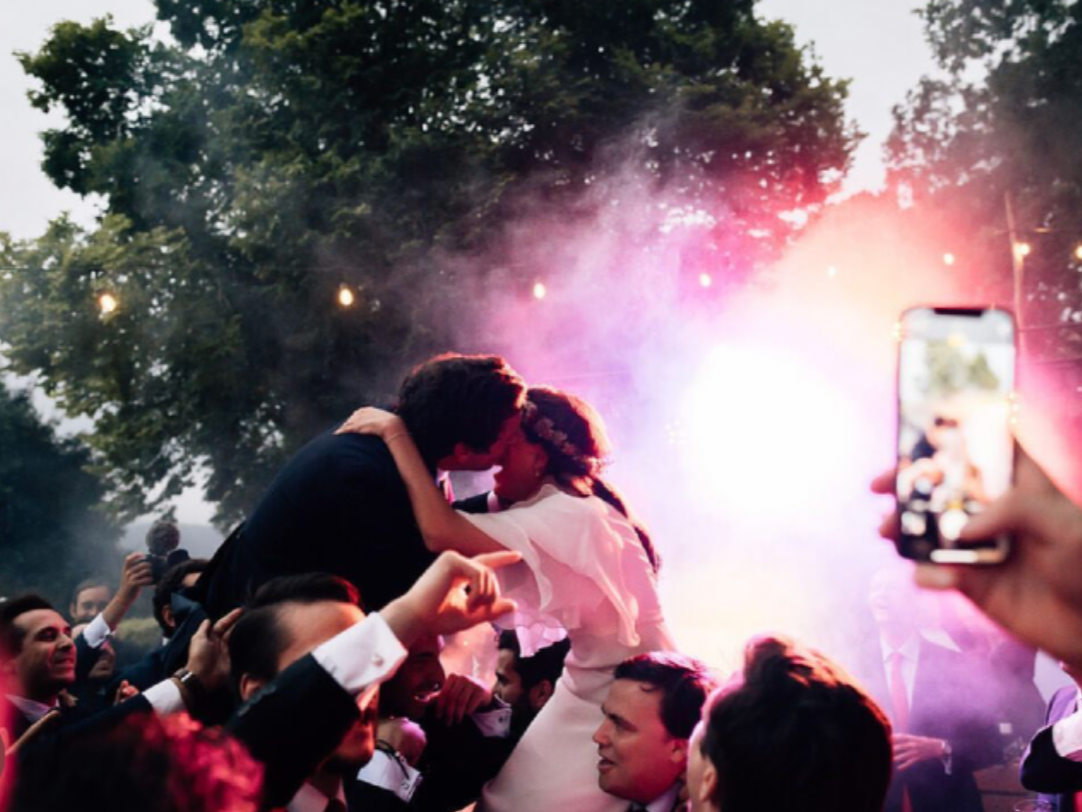 Pareja joven bailando en una fiesta al aire libre, rodeados de personas, con luces y humo en el fondo. La pareja está abrazada y levantada en el aire, en un ambiente festivo.