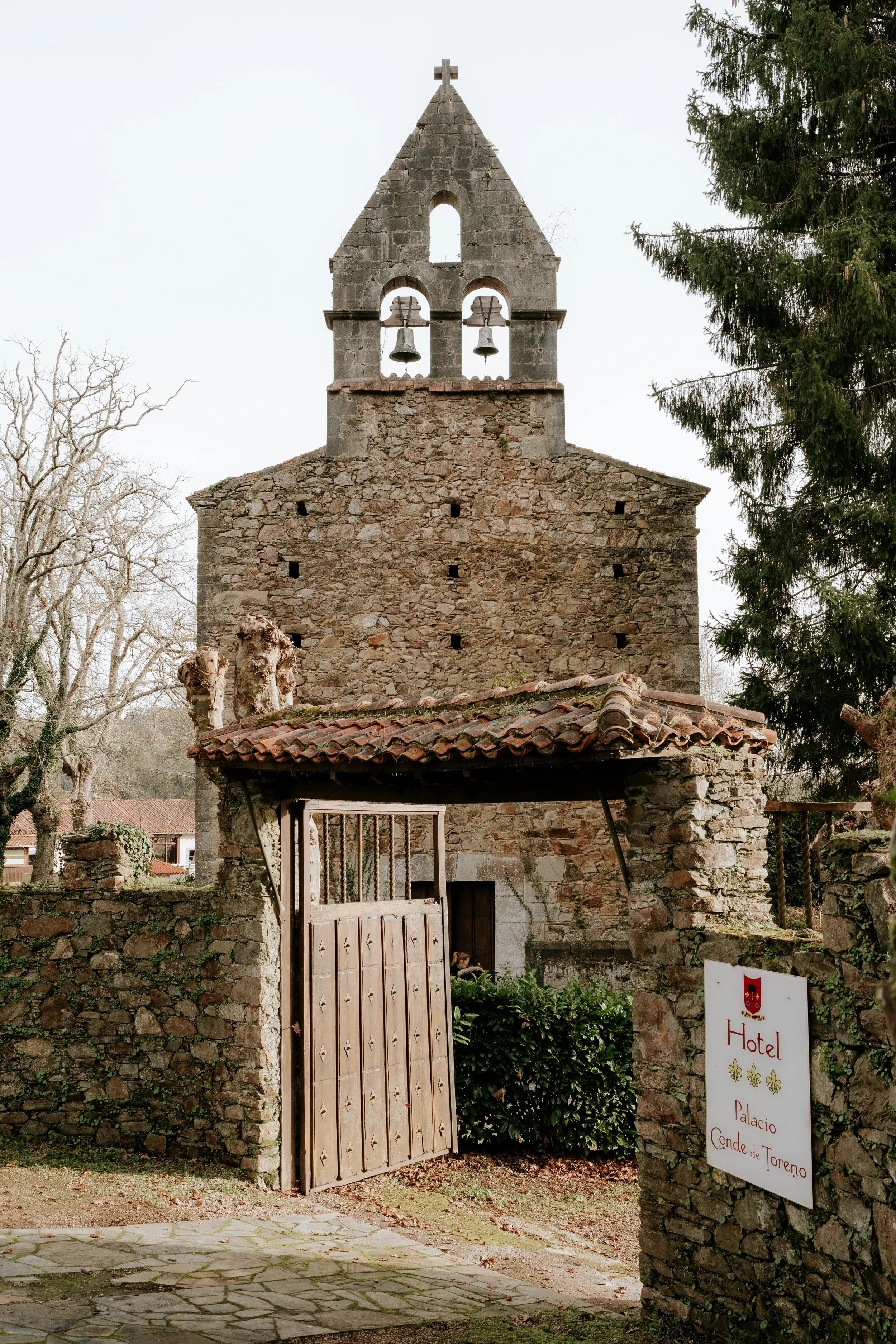 Una iglesia antigua de piedra con campanario y campanas, un portón de madera, y un cartel que dice 'Hotel Palacio Conde de Toreno'.