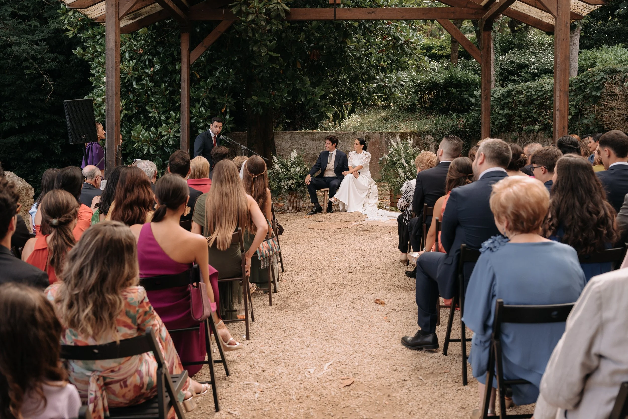 Una ceremonia de boda al aire libre con una pareja sentada en el altar, rodeada de invitados sentados en sillas, con un arco de flores y árboles verdes de fondo.