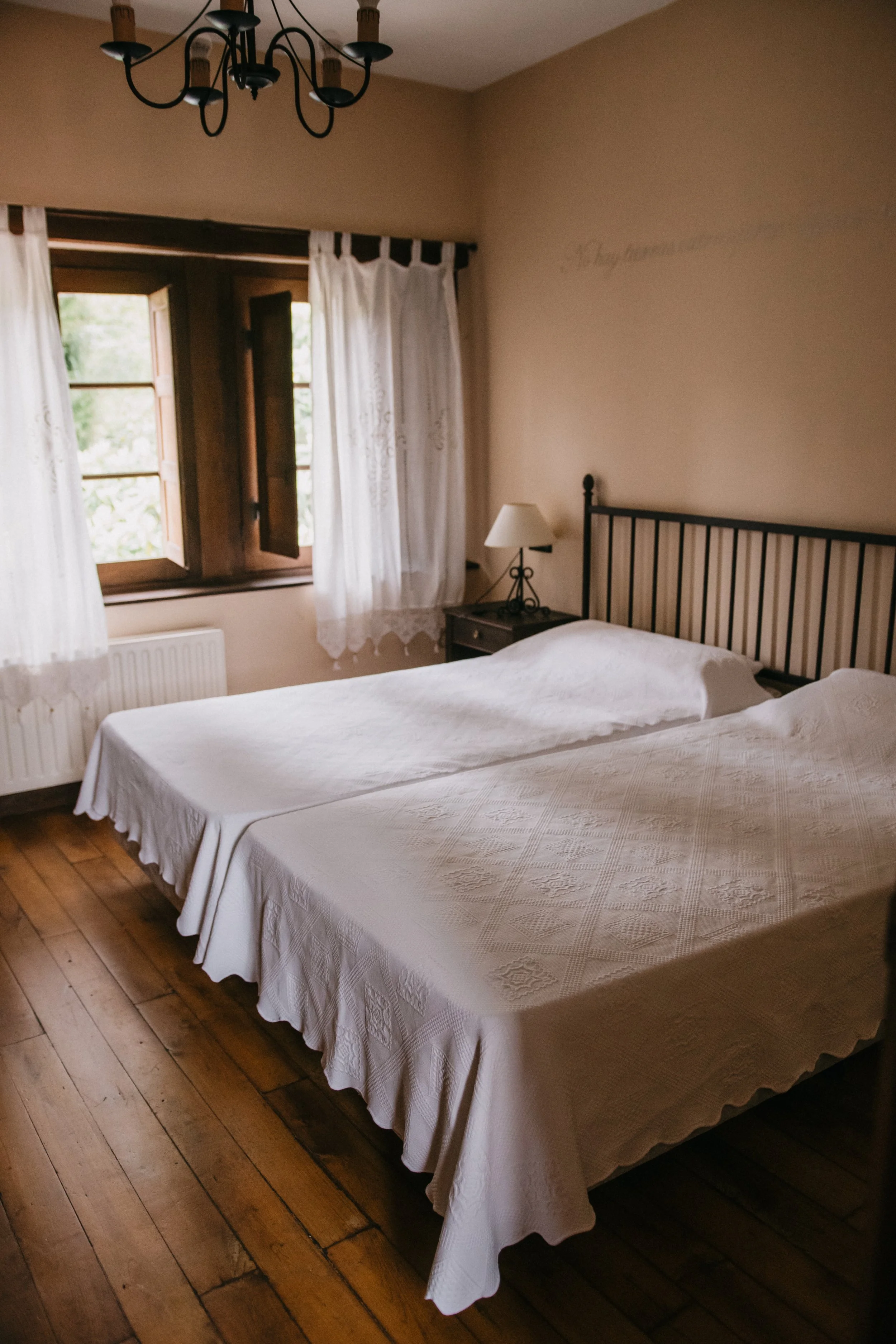 Habitación con cama doble con colcha blanca, ventanas abiertas con cortinas blancas, lámpara en la mesita de noche, decoraciones sencillas en paredes y suelo de madera.