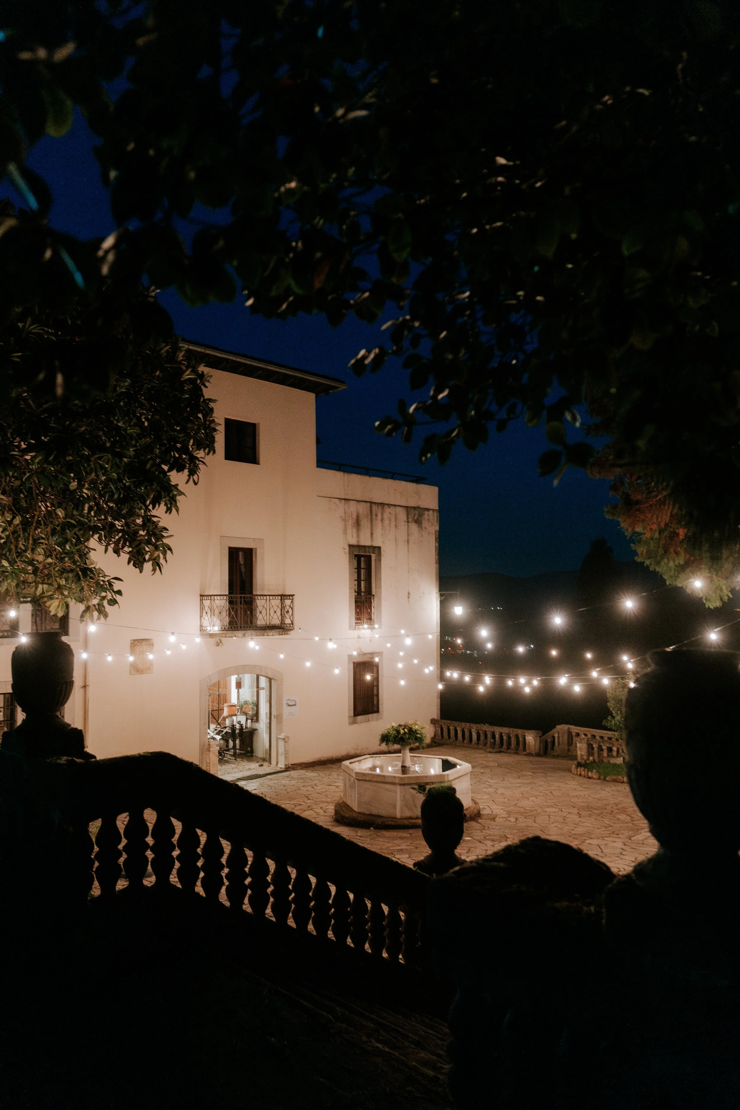 Vista nocturna de un patio con edificio blanco, decorado con luces colgantes y una fuente en el centro, rodeado de árboles y barandillas.