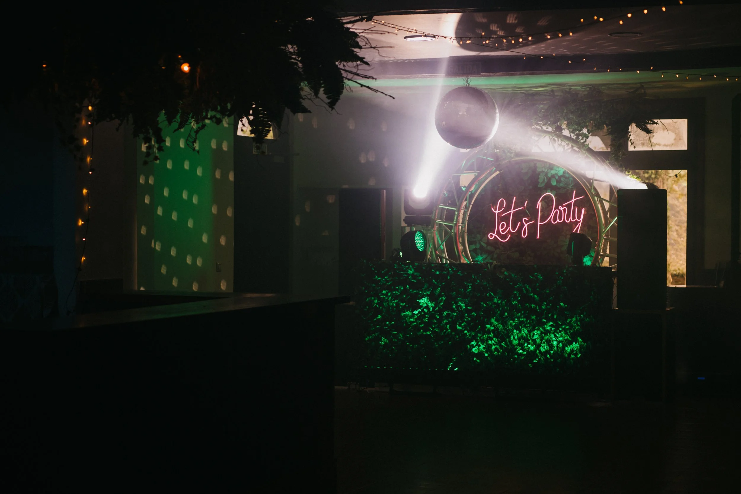 Decoración para fiesta con luces verdes y un letrero de neón que dice 'Let's Party', con una bola de discoteca y luces dirigidas hacia un escenario.