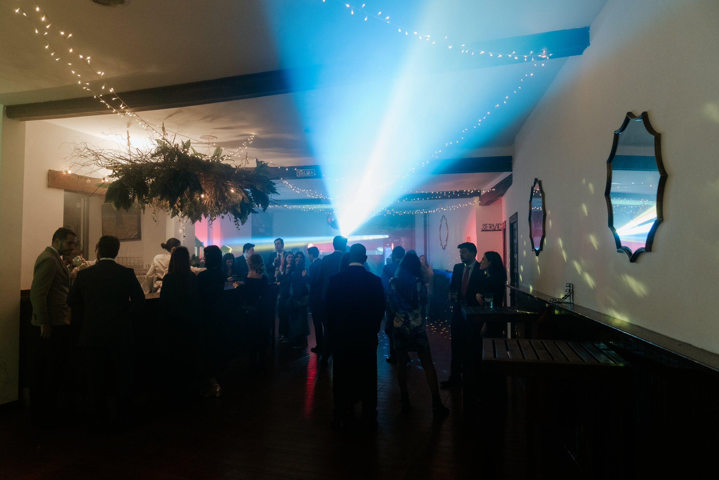 Personas en una fiesta dentro de un salón decorado con luces y un árbol colgado del techo, con efectos de iluminación en la pista de baile.
