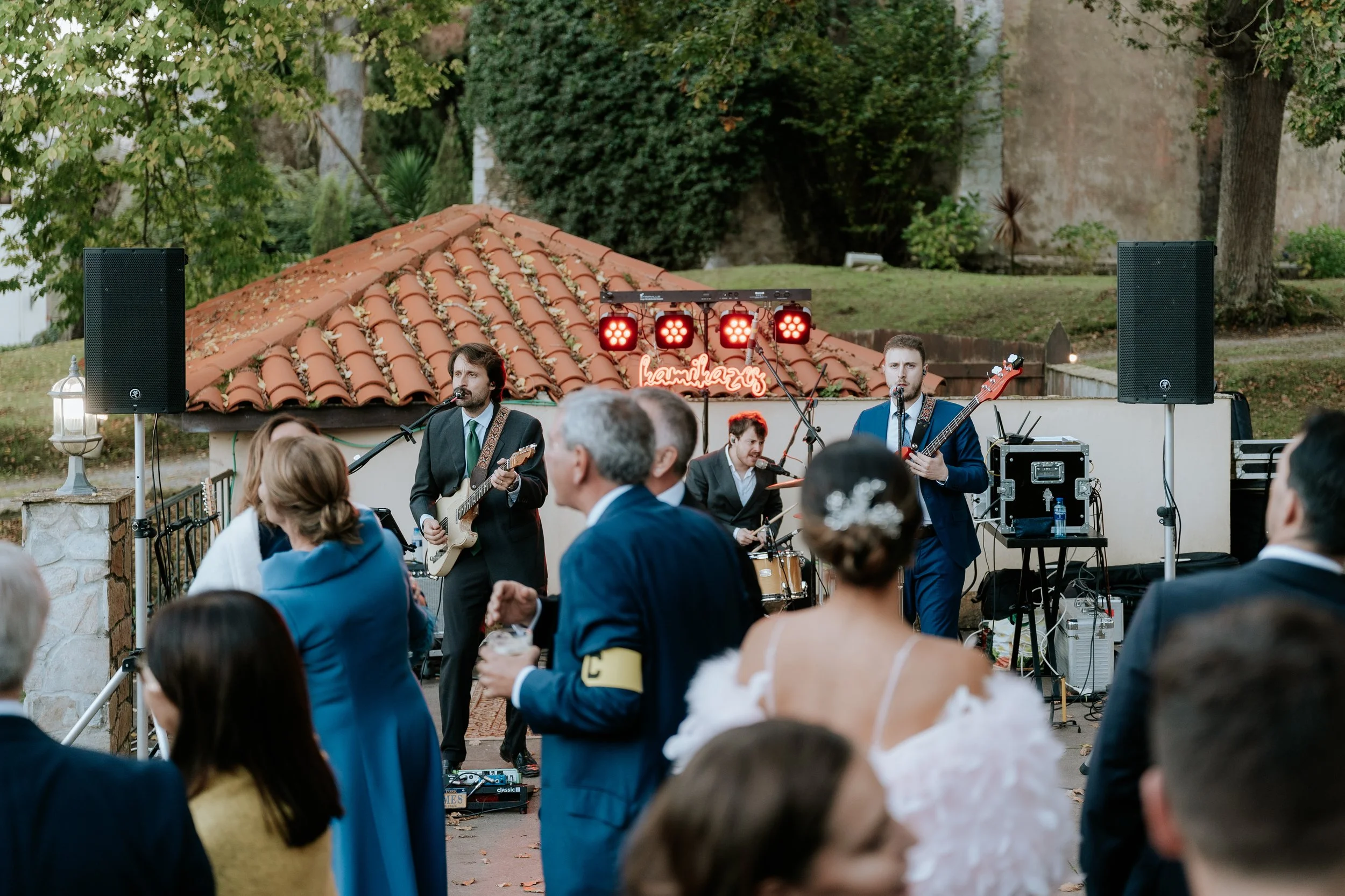Banda musical tocando en un evento al aire libre, con varias personas en el público disfrutando la música, en un entorno con árboles y un techo de tejas resaltando en el fondo.
