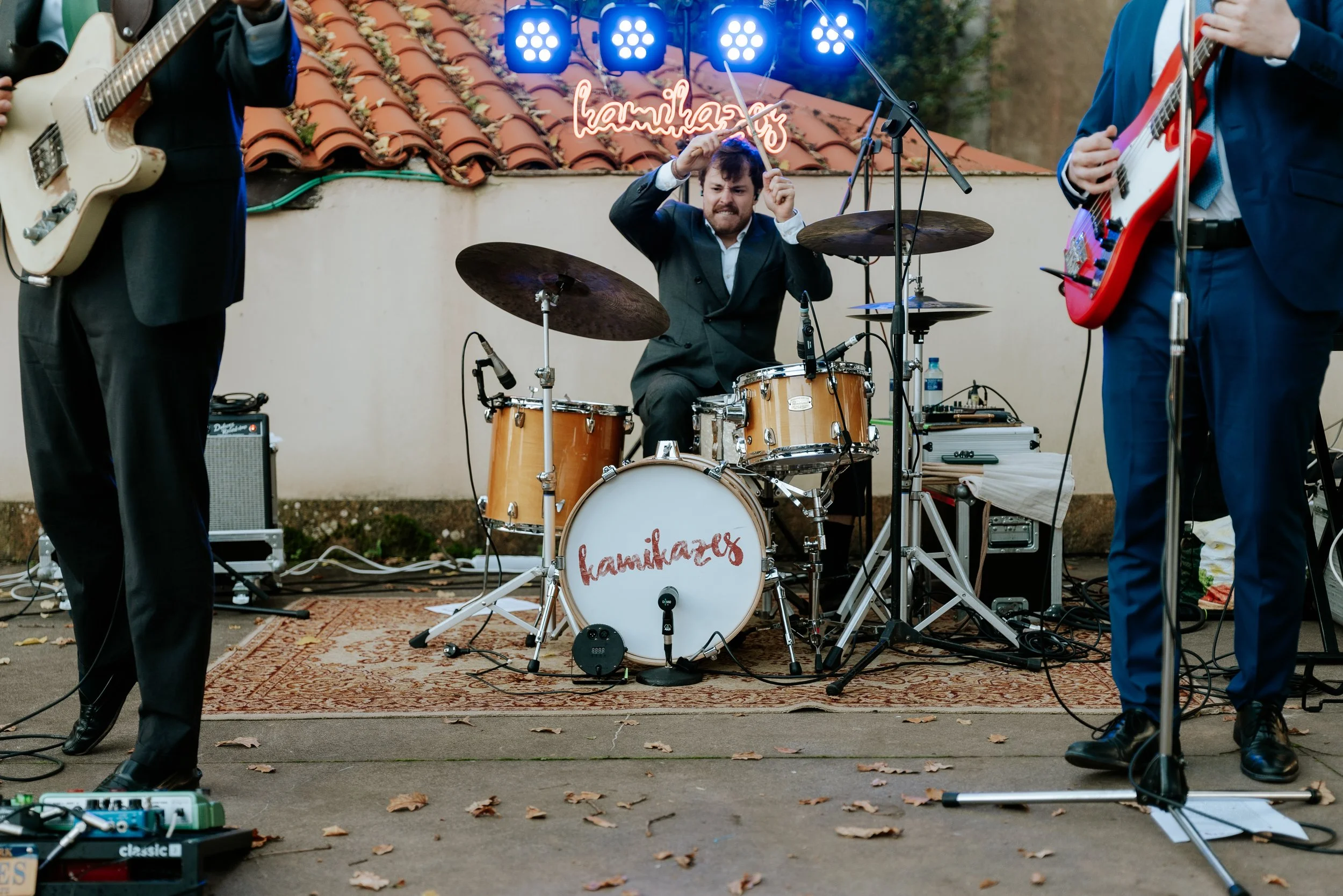 Un grupo de músicos tocando en un escenario al aire libre con un fondo de tejado y una pared, un batería en el centro y dos guitarristas a los lados. El tambor lleva el nombre 'kamikazes' y hay una luz de neón con la misma palabra.