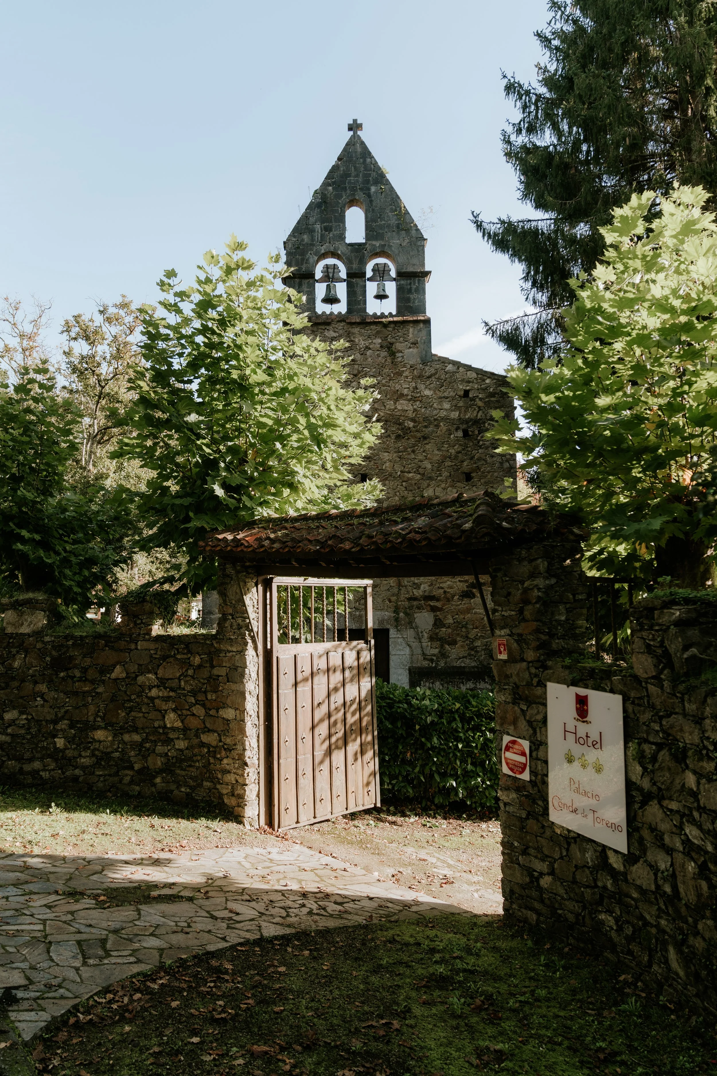 Una iglesia de piedra con campanario y reloj, rodeada de árboles y un muro de piedra, con una entrada de madera y un cartel que indica un hotel llamado 'Palacio Conde de Toreno'.