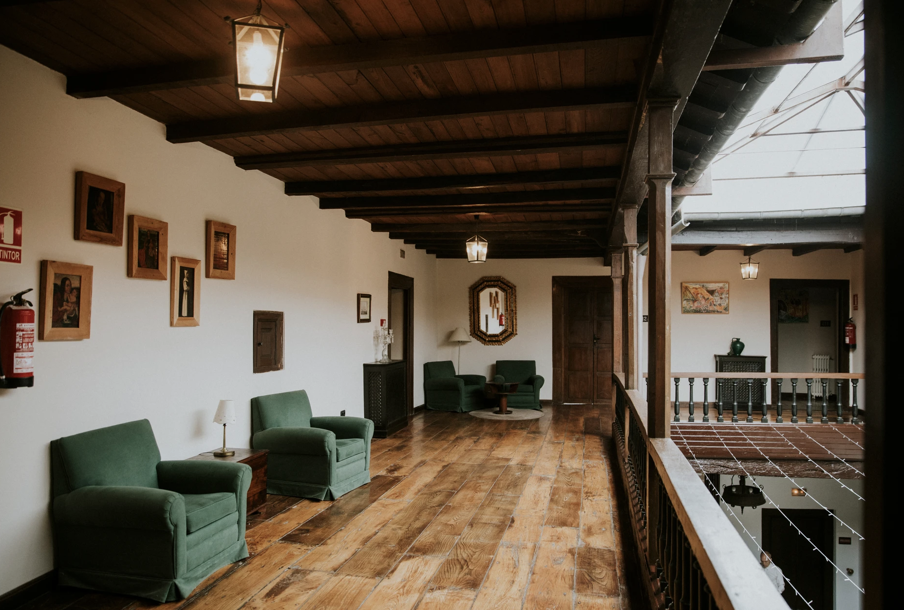 Sala de estilo rústico con sillones verdes, cuadros en la pared, lámparas de techo, piso de madera, y barandal de madera con vista al interior.