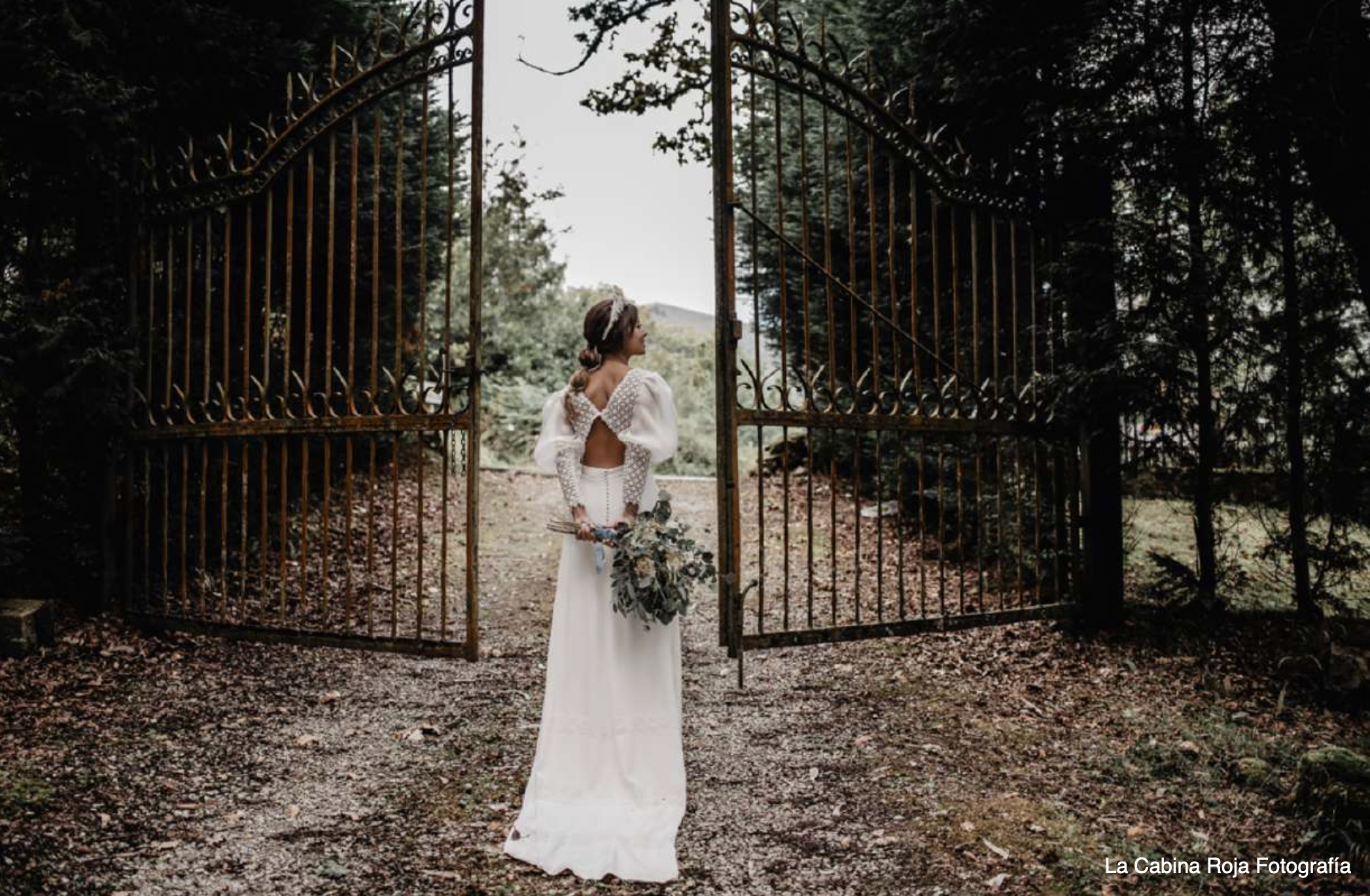 Una mujer en vestido blanco de novia sosteniendo un ramo de flores, de espaldas, de pie frente a una puerta de hierro abierta en un entorno natural rodeado de árboles.