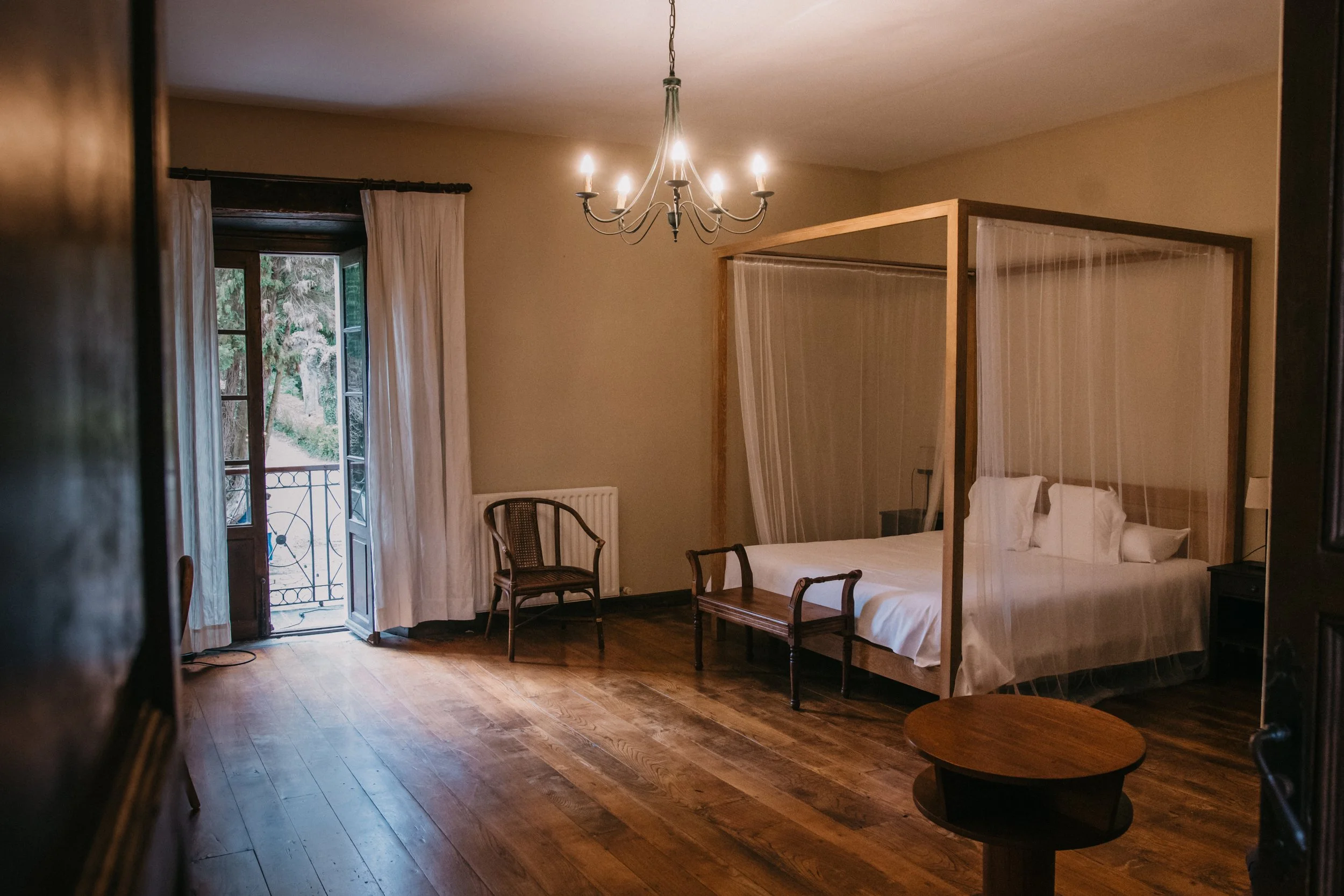 Habitación con cama de matrimonial con cortinas ligeras, sillas de madera, piso de madera, con balcón abierto y vista a la naturaleza, lámpara de techo y paredes claras.