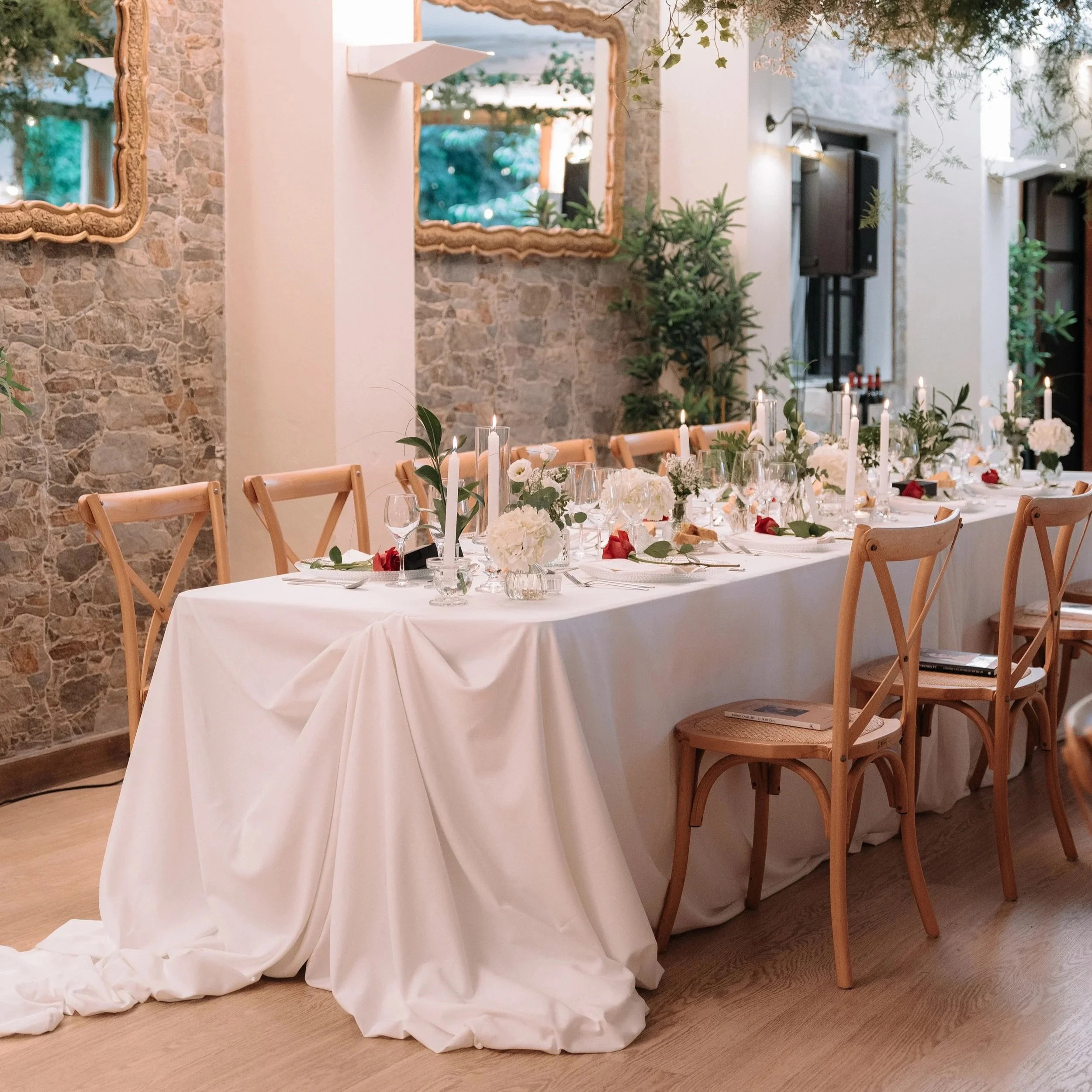 Mesa decorada para un evento en un ambiente elegante, con mantel blanco, velas y arreglos florales, y sillas de madera alrededor.