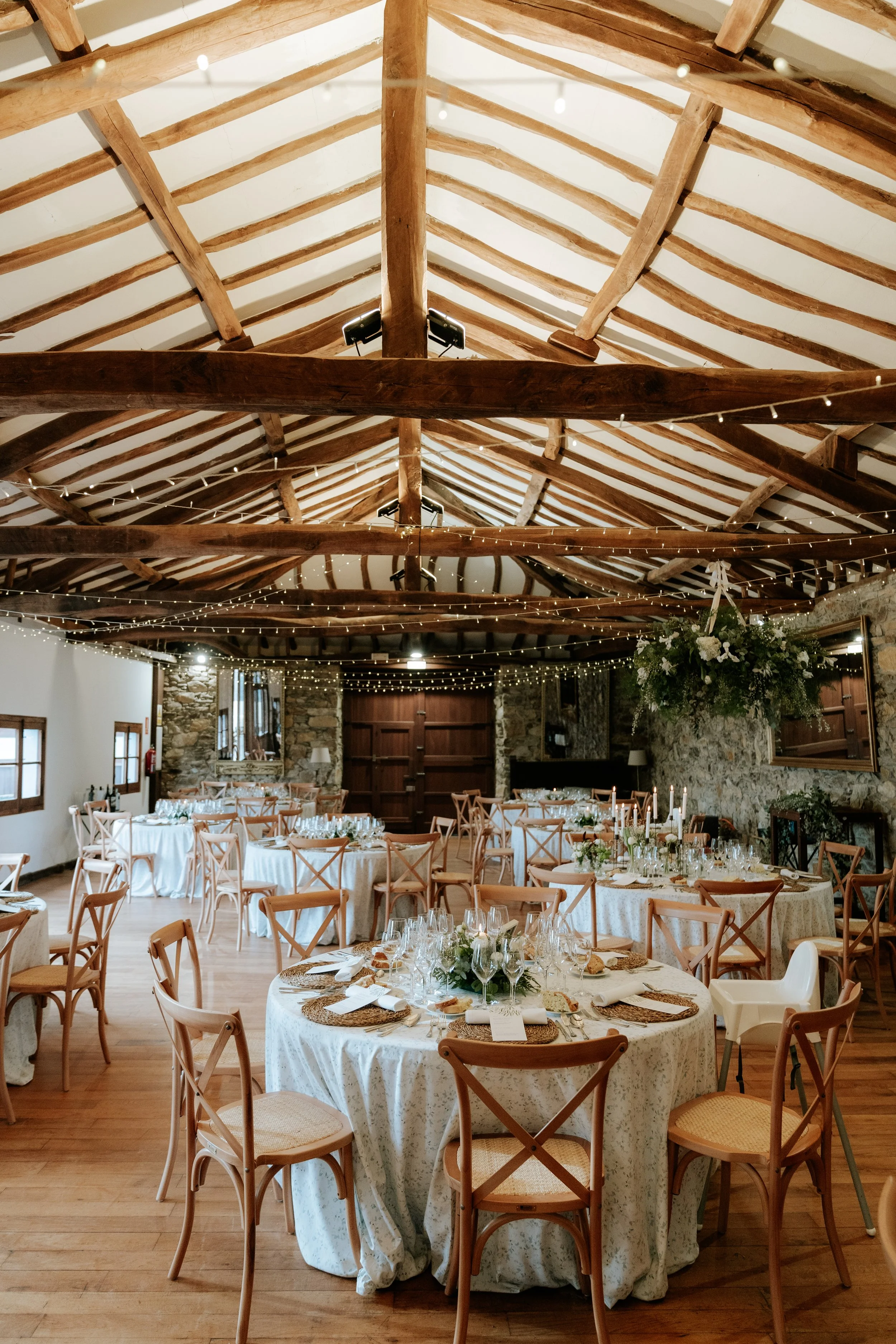 Sala de eventos decorada con mesas redondas con manteles blancos, copas y platos, con techos de madera expuesta y decoraciones con luces y flores.