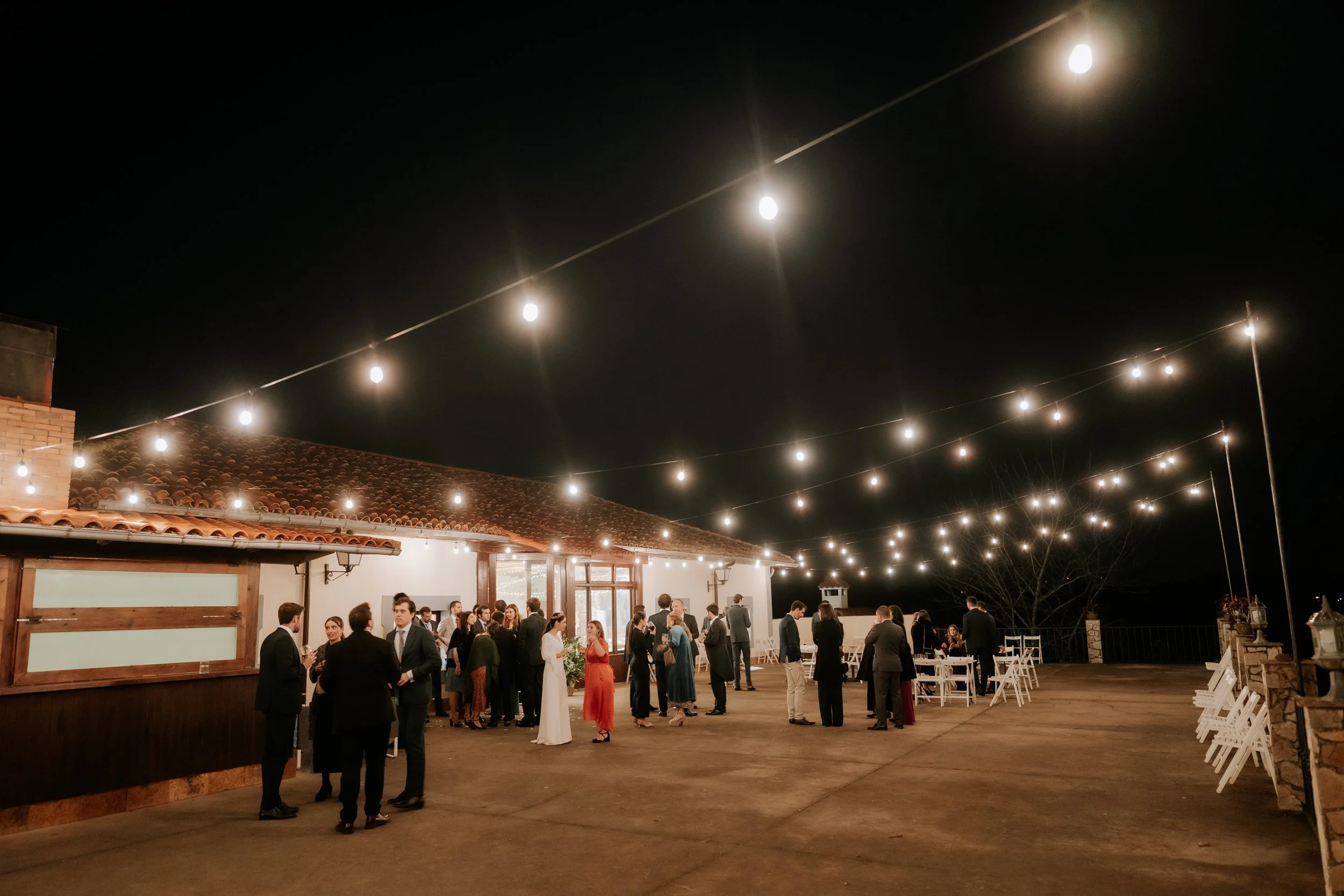 Reunión social al aire libre en la noche con luces colgantes, personas conversando y disfrutando en un espacio con estructura de tejas y ambiente festivo.