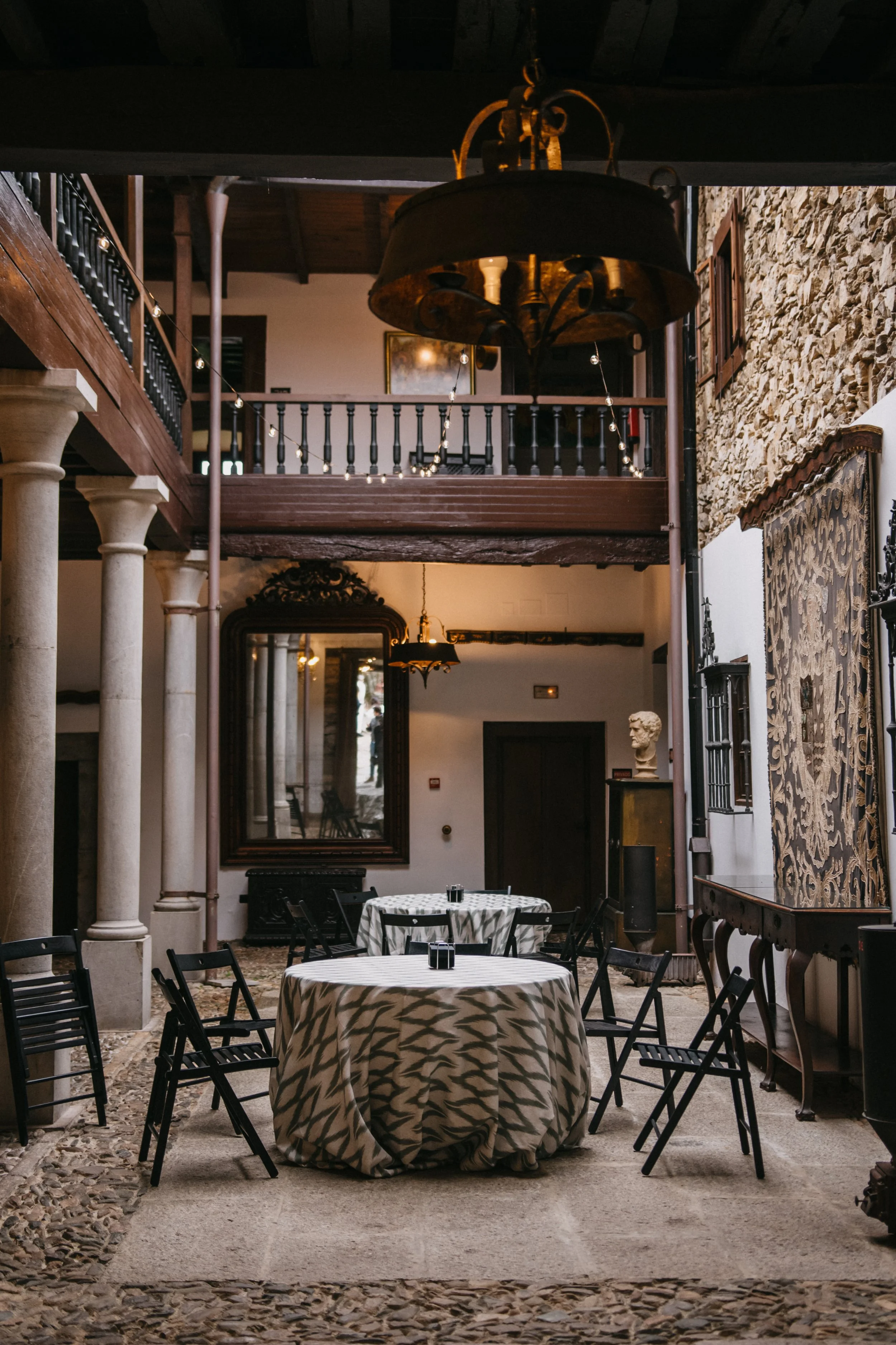 Interior de un restaurante con mesas redondas cubiertas con manteles de caimán, sillas negras, columnas de piedra, un espejo grande y decoraciones clásicas.